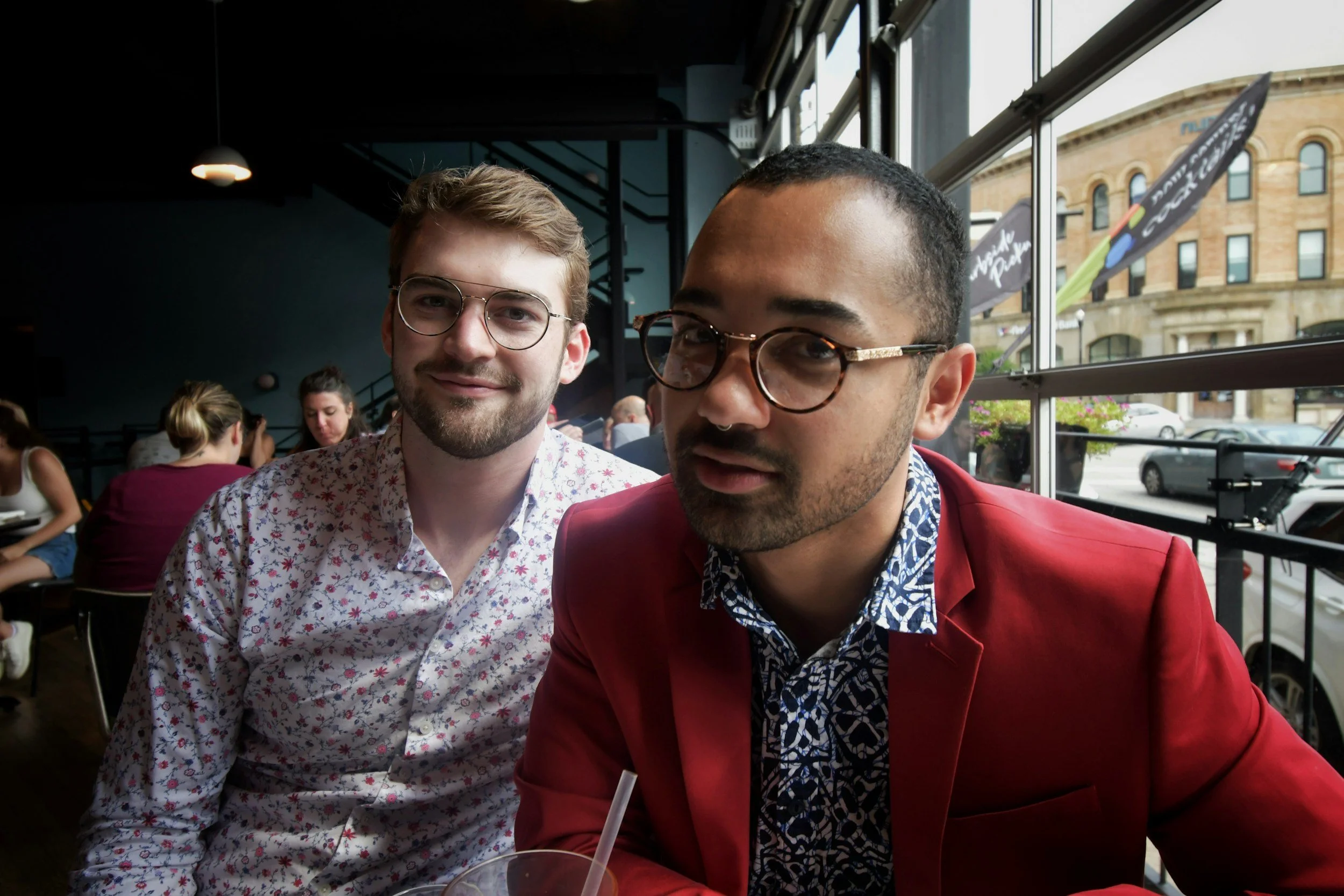 Two men sitting at a restaurant table near a window, with other diners in the background. One man has glasses, a beard, and wearing a floral shirt. The other man has short hair, glasses, and is wearing a red blazer with a patterned shirt underneath.
