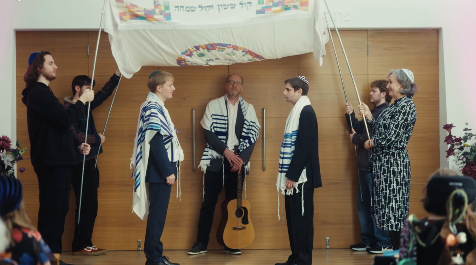 People participating in a Jewish ceremony under a canopy, with men wearing prayer shawls and kippahs, and a woman holding the canopy poles, while a man with a guitar stands behind them.