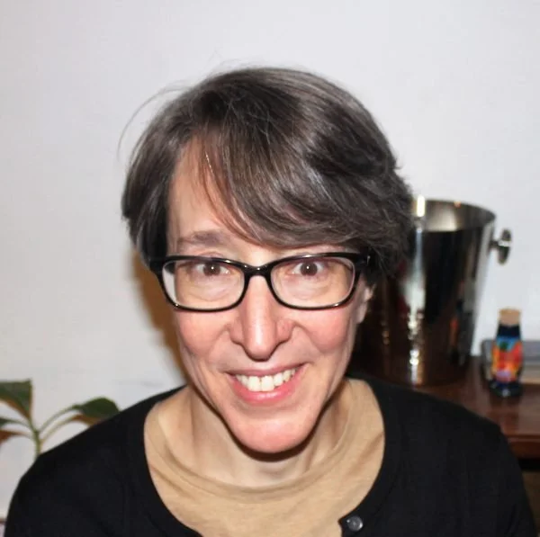 A woman with short brown hair, wearing glasses and a black shirt, smiling in front of a white wall and a wooden table with a silver ice bucket and a small colorful object.