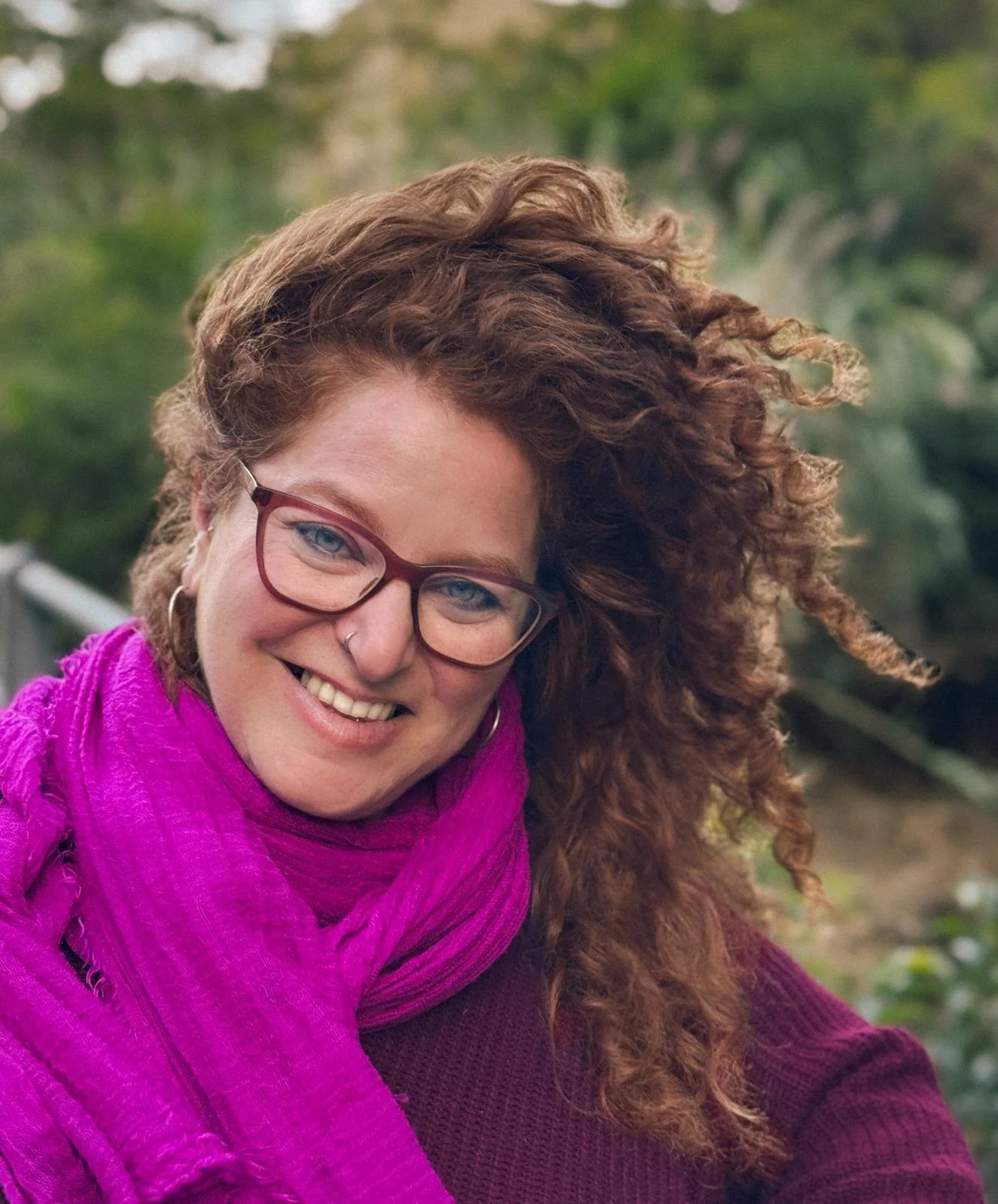 A woman with curly red hair, glasses, and a vibrant pink scarf smiling outdoors with a blurred green background.