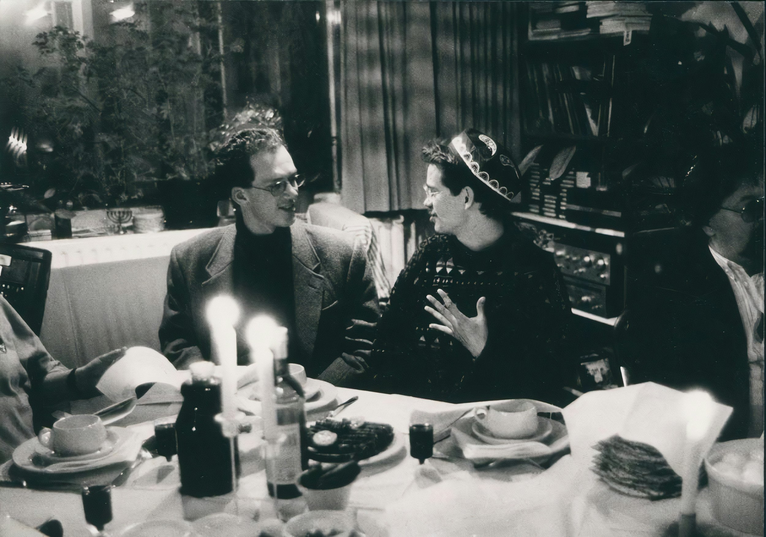 Black and white photo of two people at a dinner table, having a conversation. The woman is wearing a head covering, and the man is wearing glasses and a blazer. The table has plates, glasses, candles, and food, with bookshelves in the background.