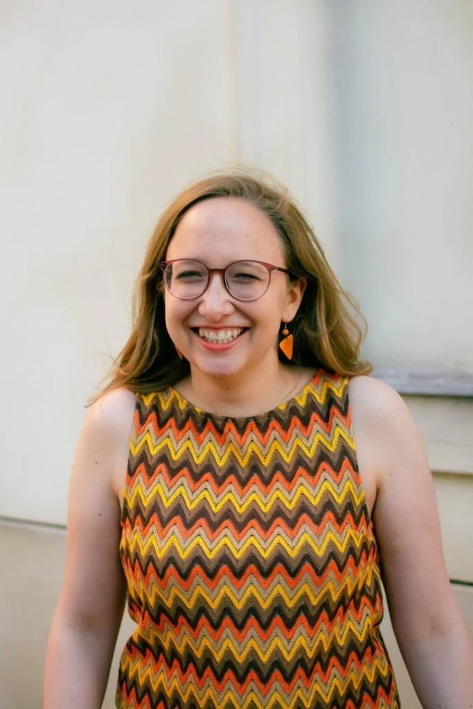 A woman with light skin, red hair, and glasses smiling outdoors in front of a neutral background. She is wearing a sleeveless dress with a zigzag pattern in orange, yellow, black, and brown.