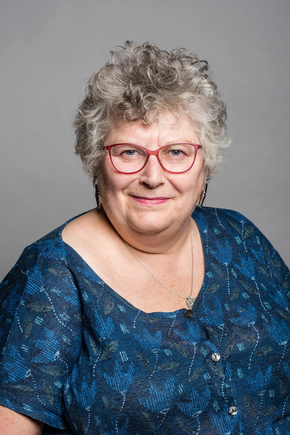 Portrait of an older woman with curly gray hair, pink glasses, wearing a blue patterned blouse, silver necklace, and earrings, against a gray background.