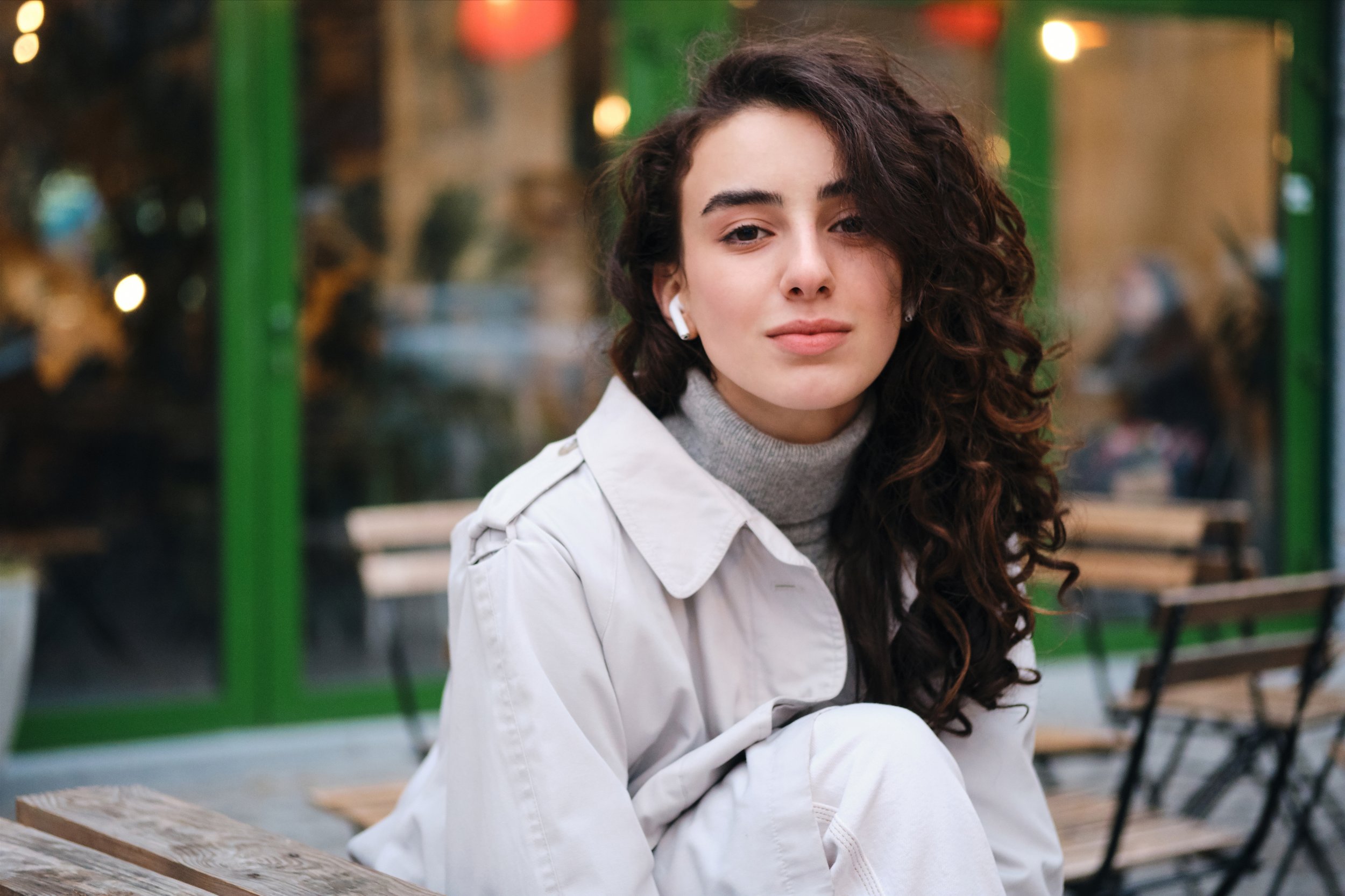 A young woman with curly brown hair, wearing a gray turtleneck and a light-colored jacket, sitting on a wooden bench outdoors near a cafe with a green window frame, during evening with blurred lights in the background.