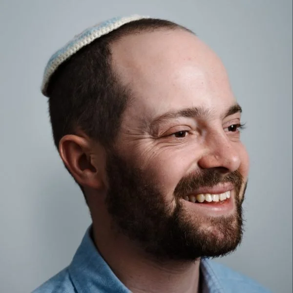 A smiling man with a beard, wearing a kippah and a denim shirt, against a gray background.