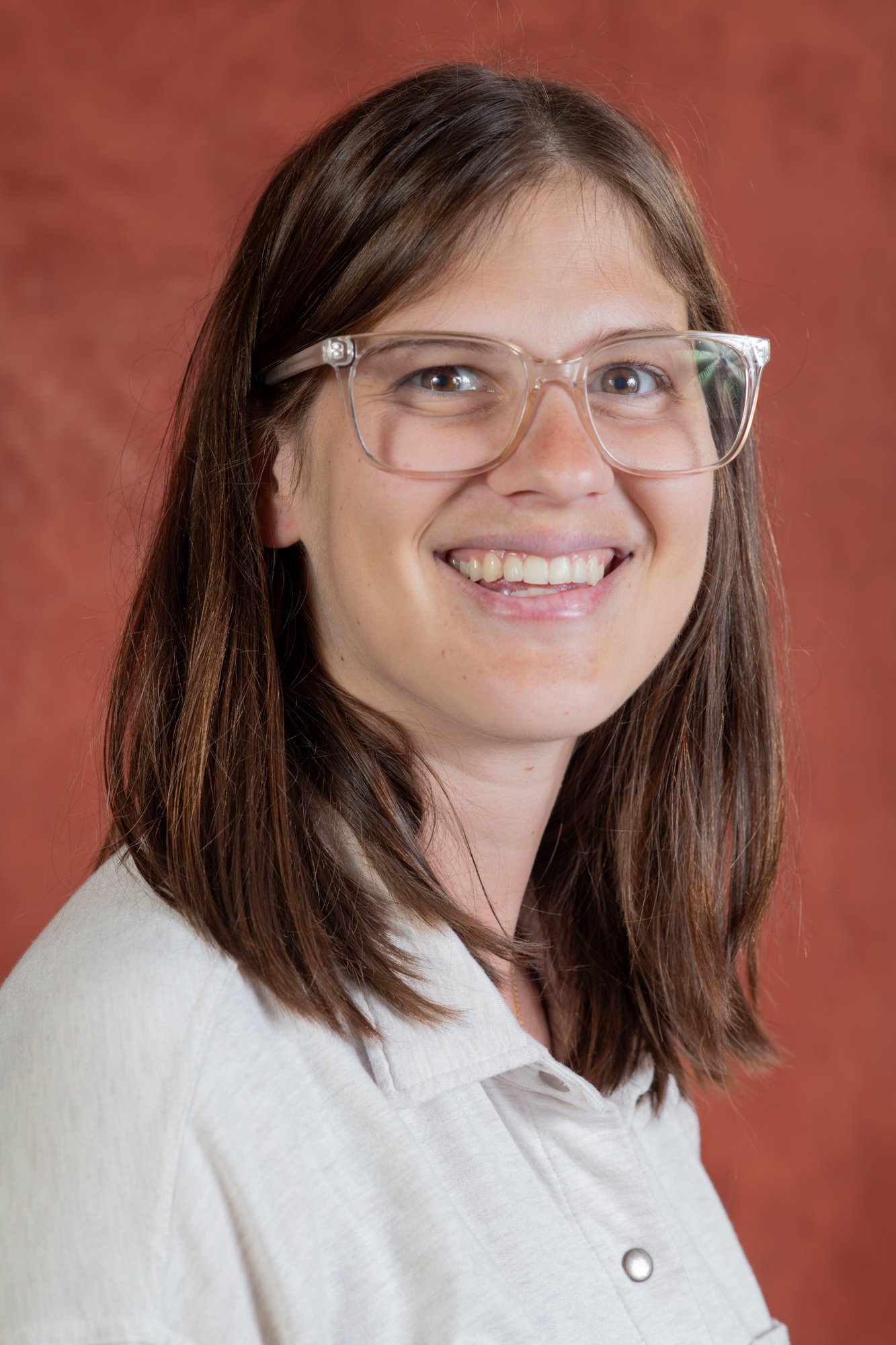Portrait of a young woman with light skin, brown hair, wearing clear glasses and a white shirt, smiling, against a reddish-brown background.