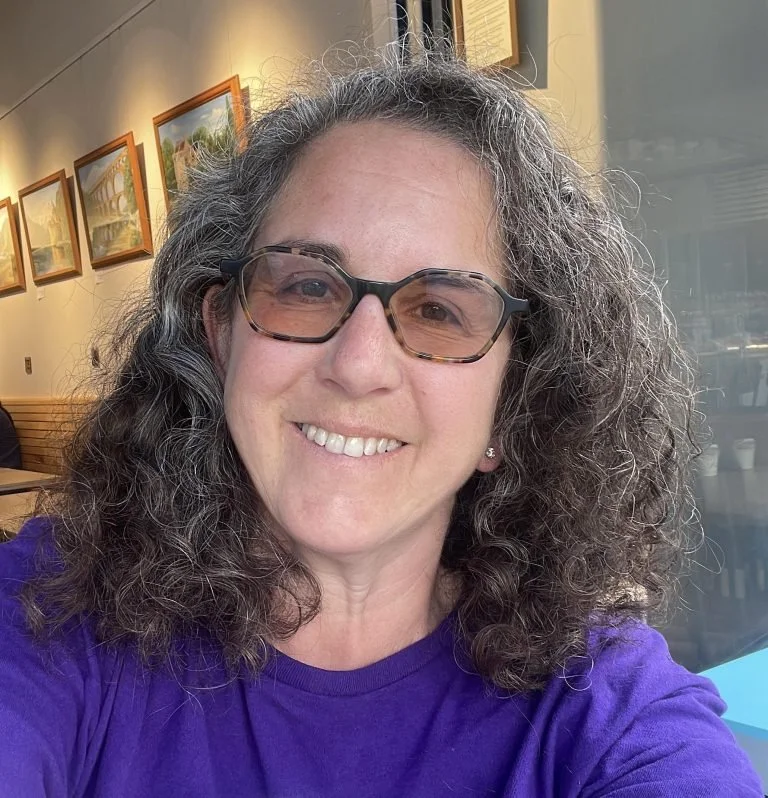 A woman with curly gray hair, glasses, and a purple shirt smiling in a cafe with framed artwork on the wall behind her.