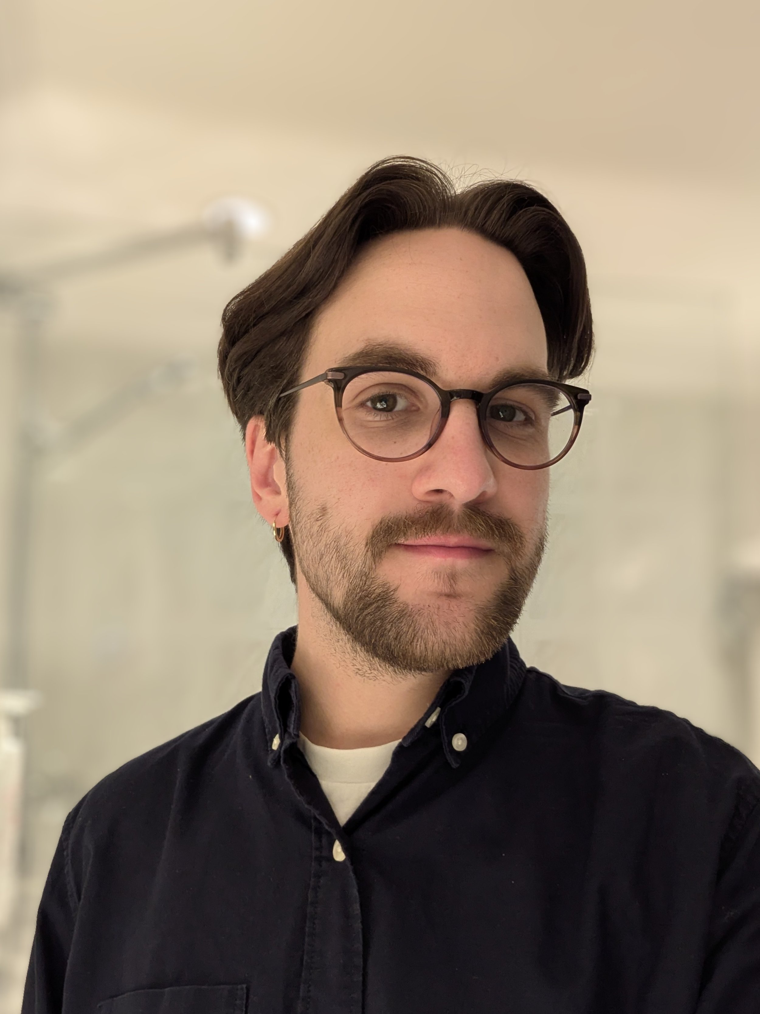 A man with dark brown hair, glasses, and a beard taking a selfie indoors, wearing a black button-up shirt and a white undershirt, with a blurry shower or curtain in the background.