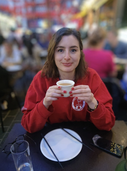 A young woman with brown hair and red lipstick, wearing a red sweatshirt, sits at a table in a busy outdoor restaurant, holding a white cup of tea or coffee.