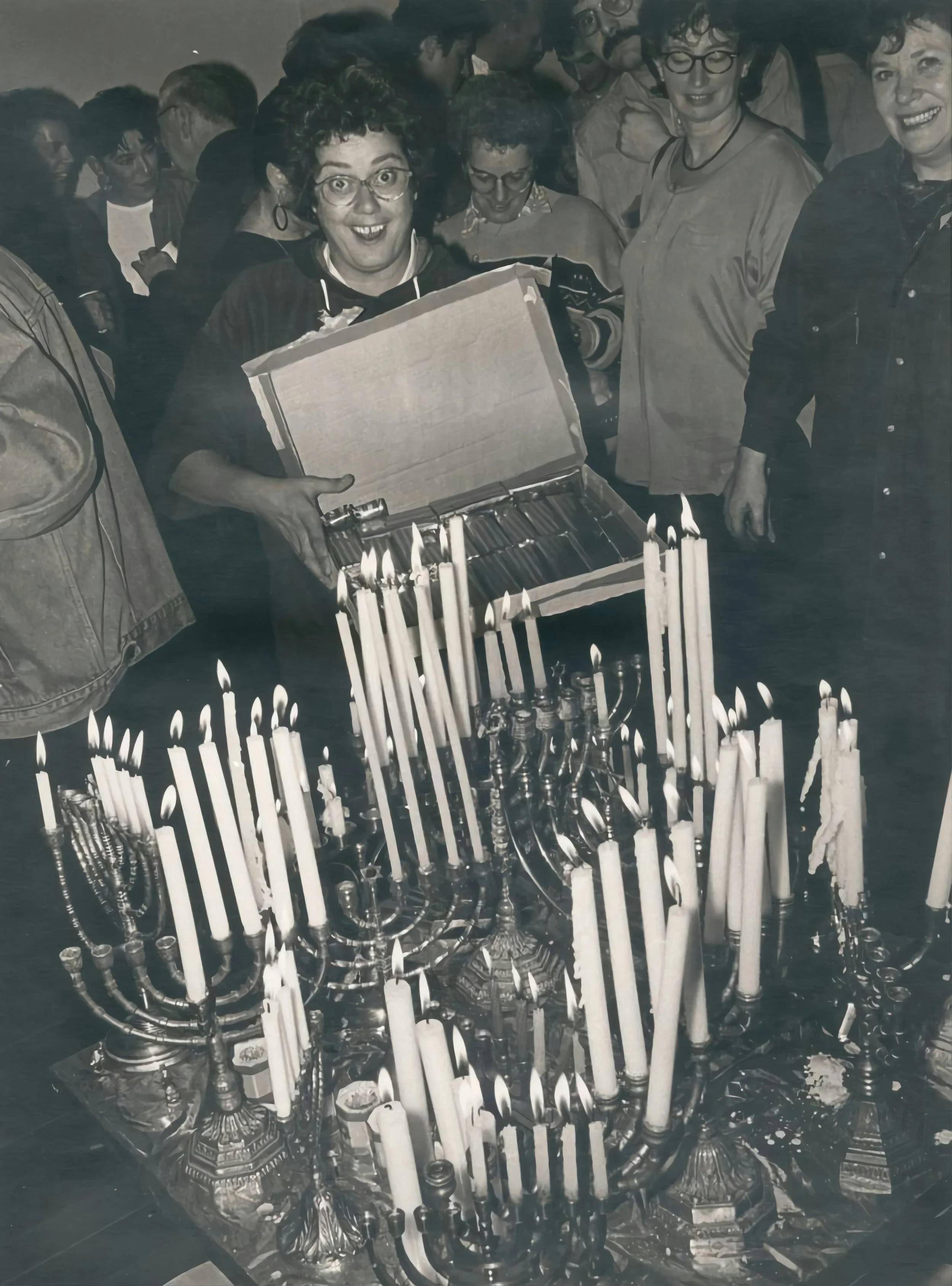 A group of women gathered around a table with lit candles, one woman holding an open box with more candles, smiling and celebrating.