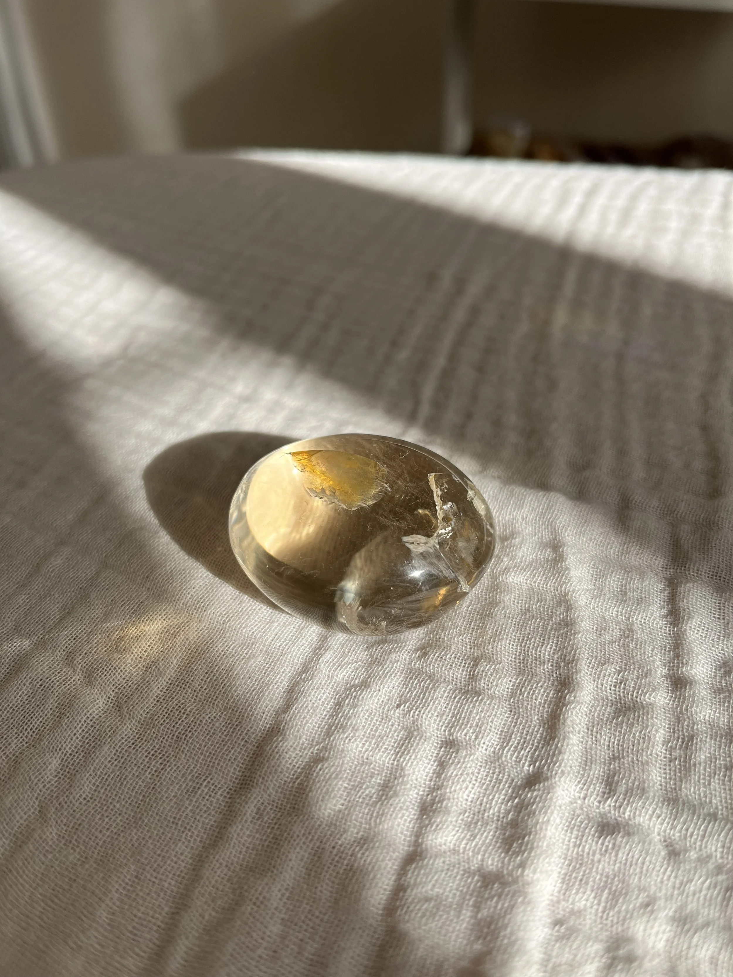 A clear, round, polished healing crystal  used for distance healing with golden streaks on a white fabric surface, sunlight casting shadows.