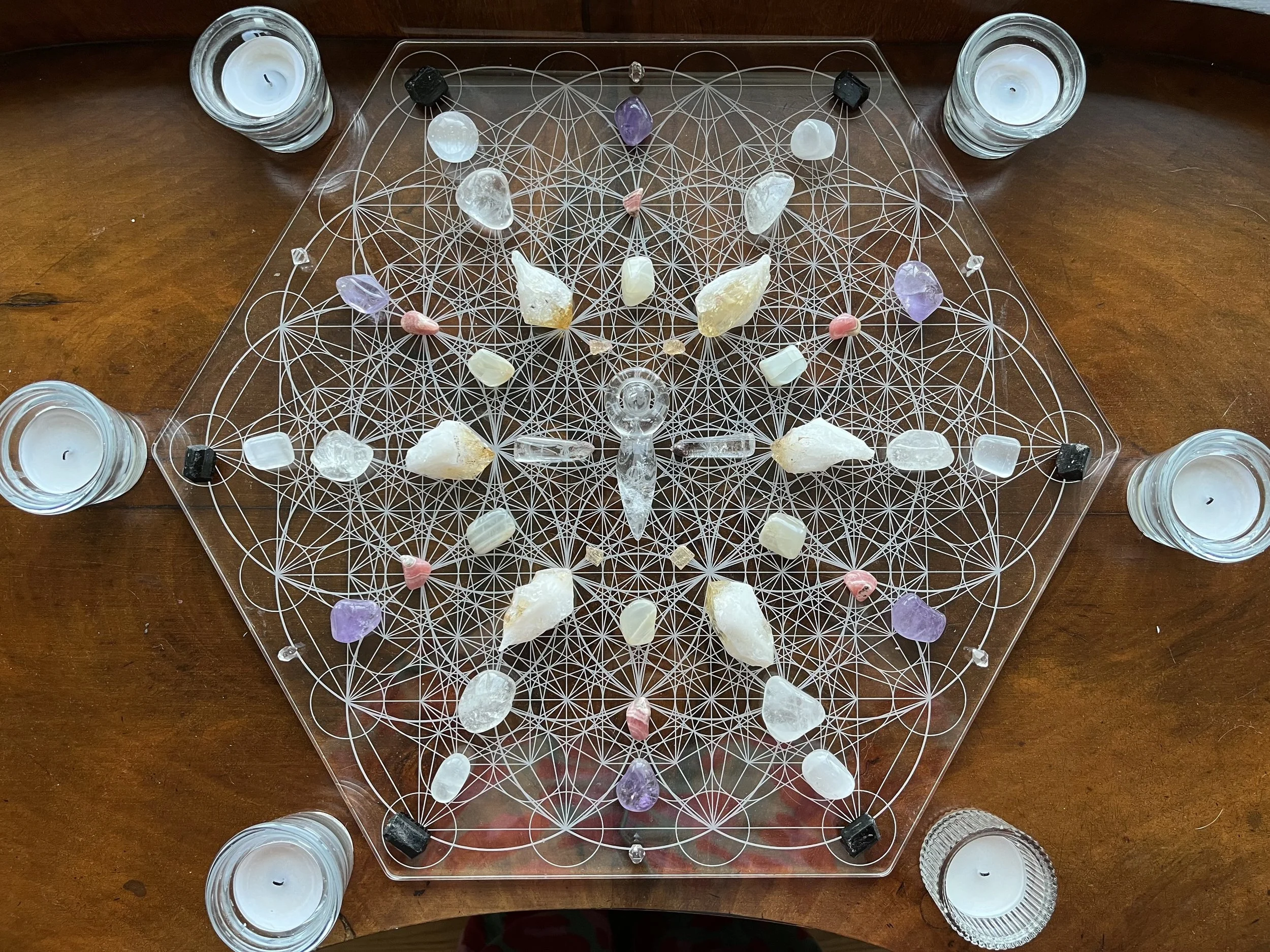 A geometric healing crystal grid with various small crystals and stones arranged on a clear acrylic surface, surrounded by five tea light candles in glass holders.