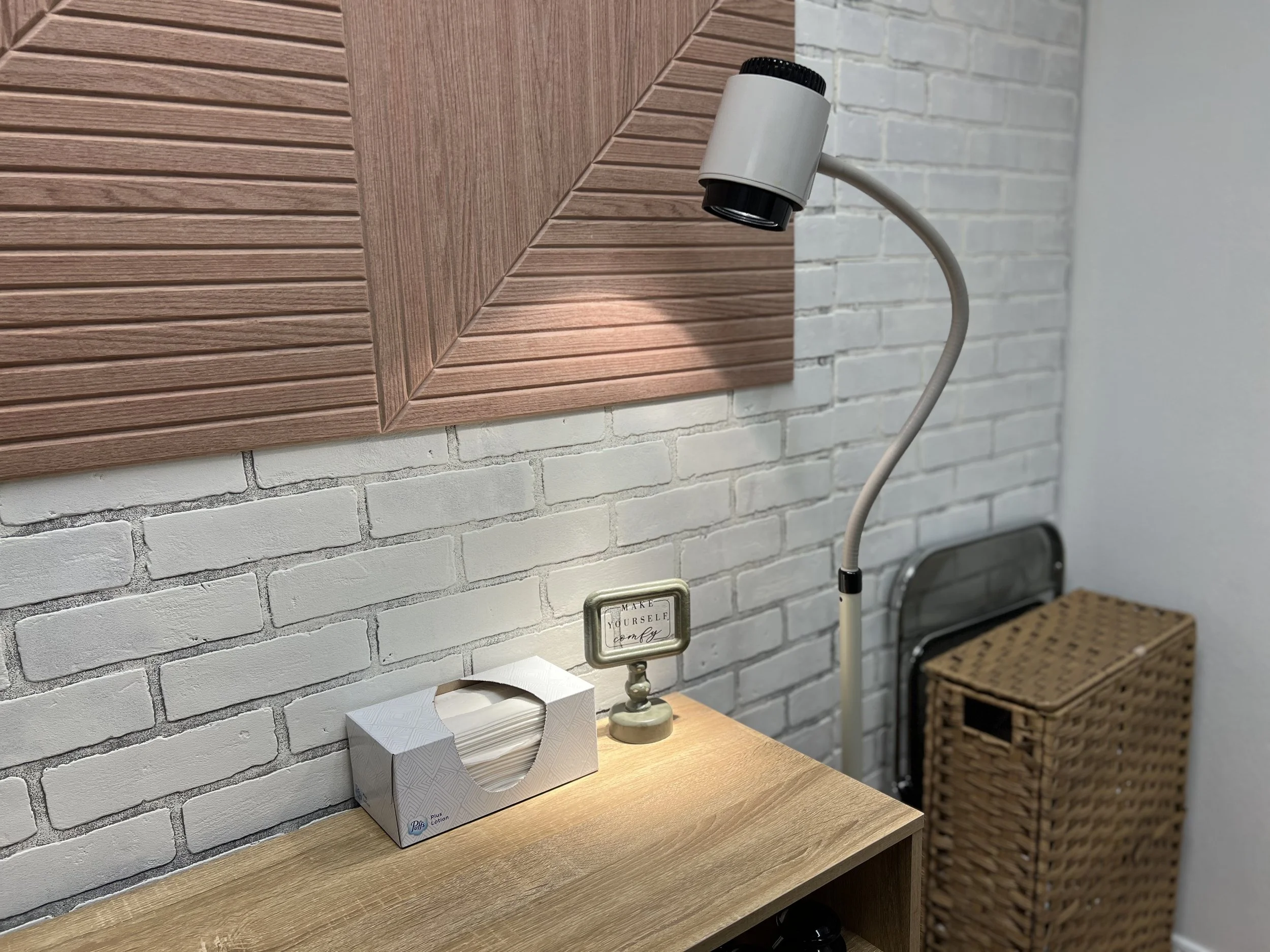 A bedside table with a tissue box, a small decorative sign, and a flexible reading lamp. Behind the table, there is a white brick wall and a wooden panel with angled slats. A wicker laundry basket is in the corner.