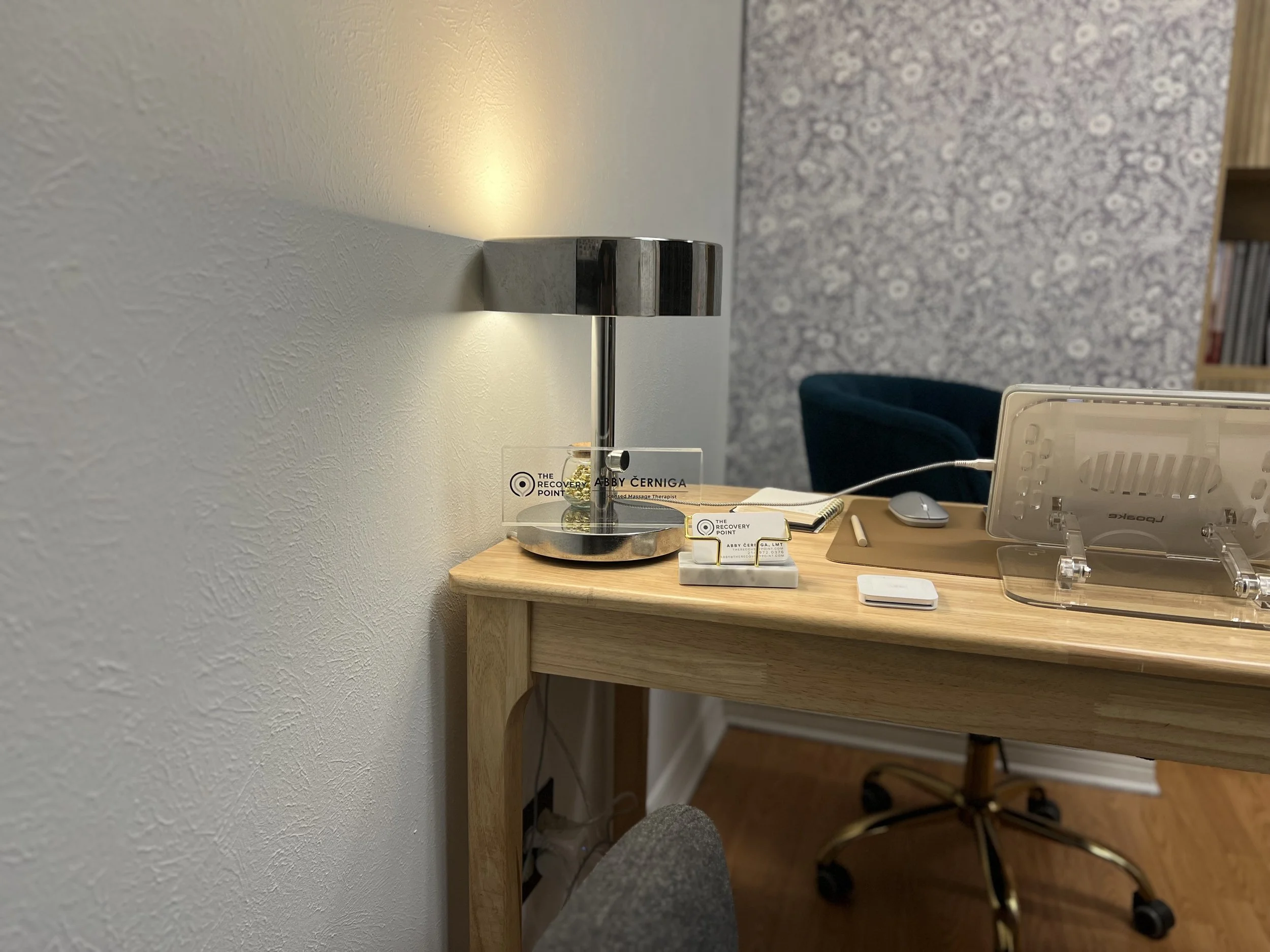 A modern wooden desk with a silver desk lamp, business cards, a mouse, a notebook, and a pen. A blue chair is visible in the background in a room with textured wallpaper.