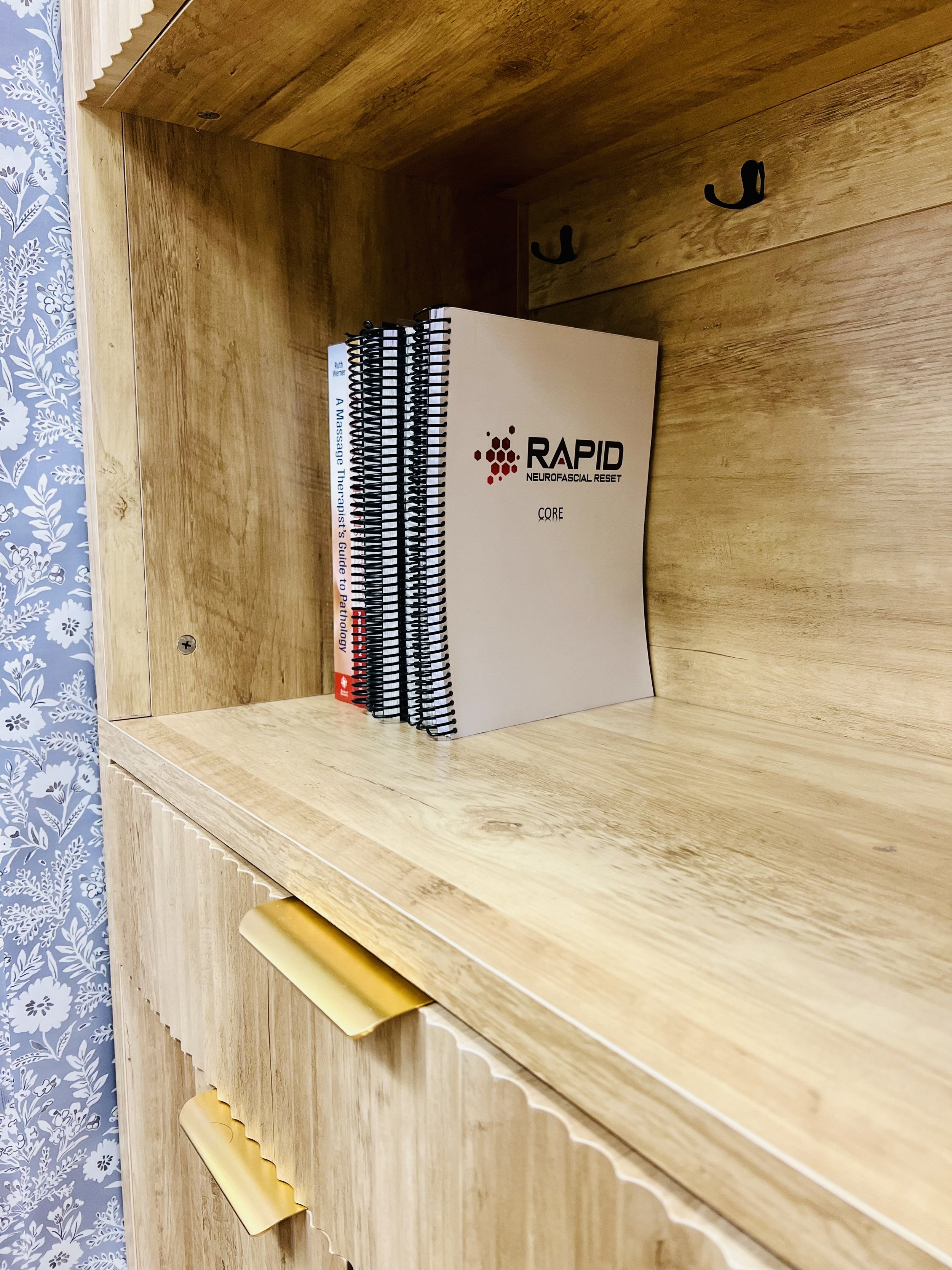 Wooden bookshelf or cabinet with books, including a spiral-bound guide titled "A Massage Therapist's Guide to Pathology" and a white manual titled "RAPID Neurofascial Reset CORE." The bookshelf has gold accents on the drawer handles and black hooks on the side.