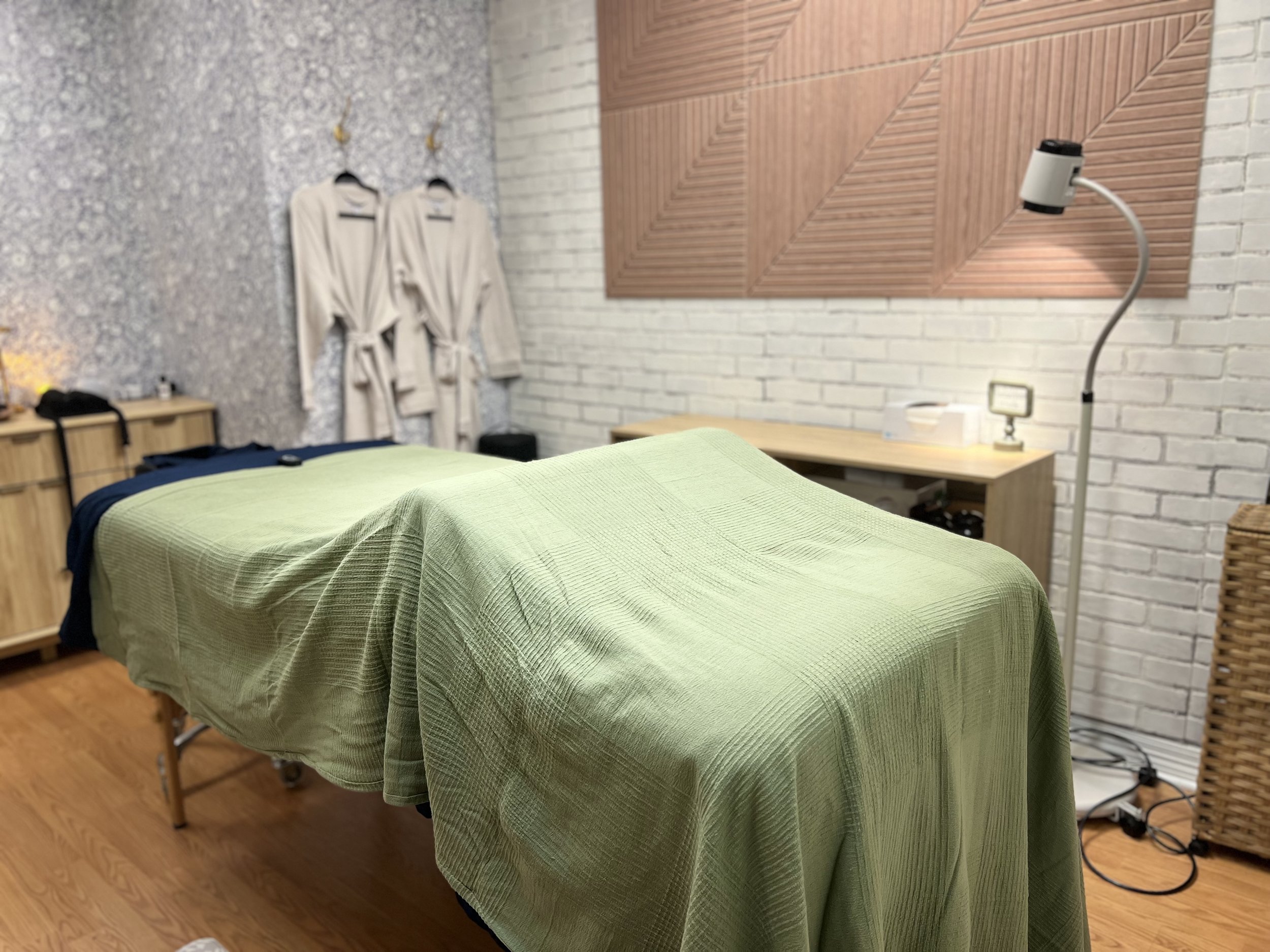 A massage table with a green cover in a room with a textured grey wall. Two white bathrobes hang on hooks on the wall. A wooden sideboard with various items, including a lamp, is in the background. There is a textured white brick wall and a wooden panel design on another wall. A tall adjustable lamp is nearby, and a wicker basket is on the right side of the room.
