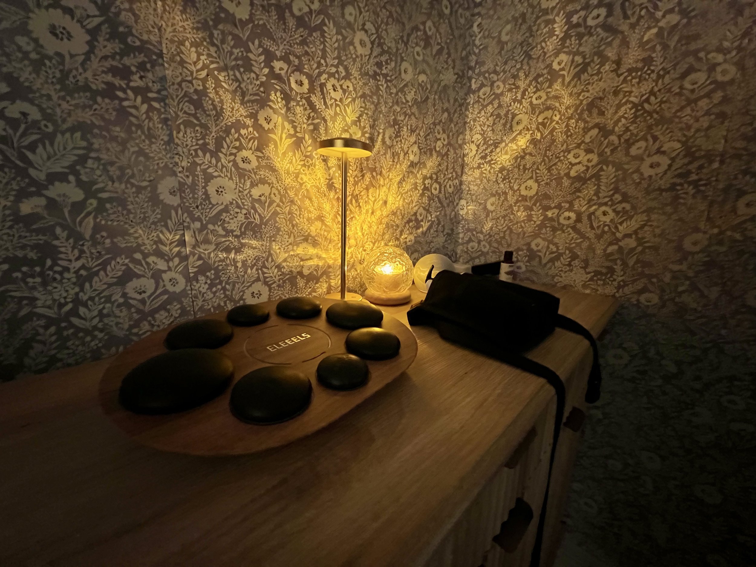 A dimly lit wooden table with a lamp casting a warm glow, a black camera bag, and a wooden plate with black stones labeled 'ELEELE' surrounded by patterned wallpaper.