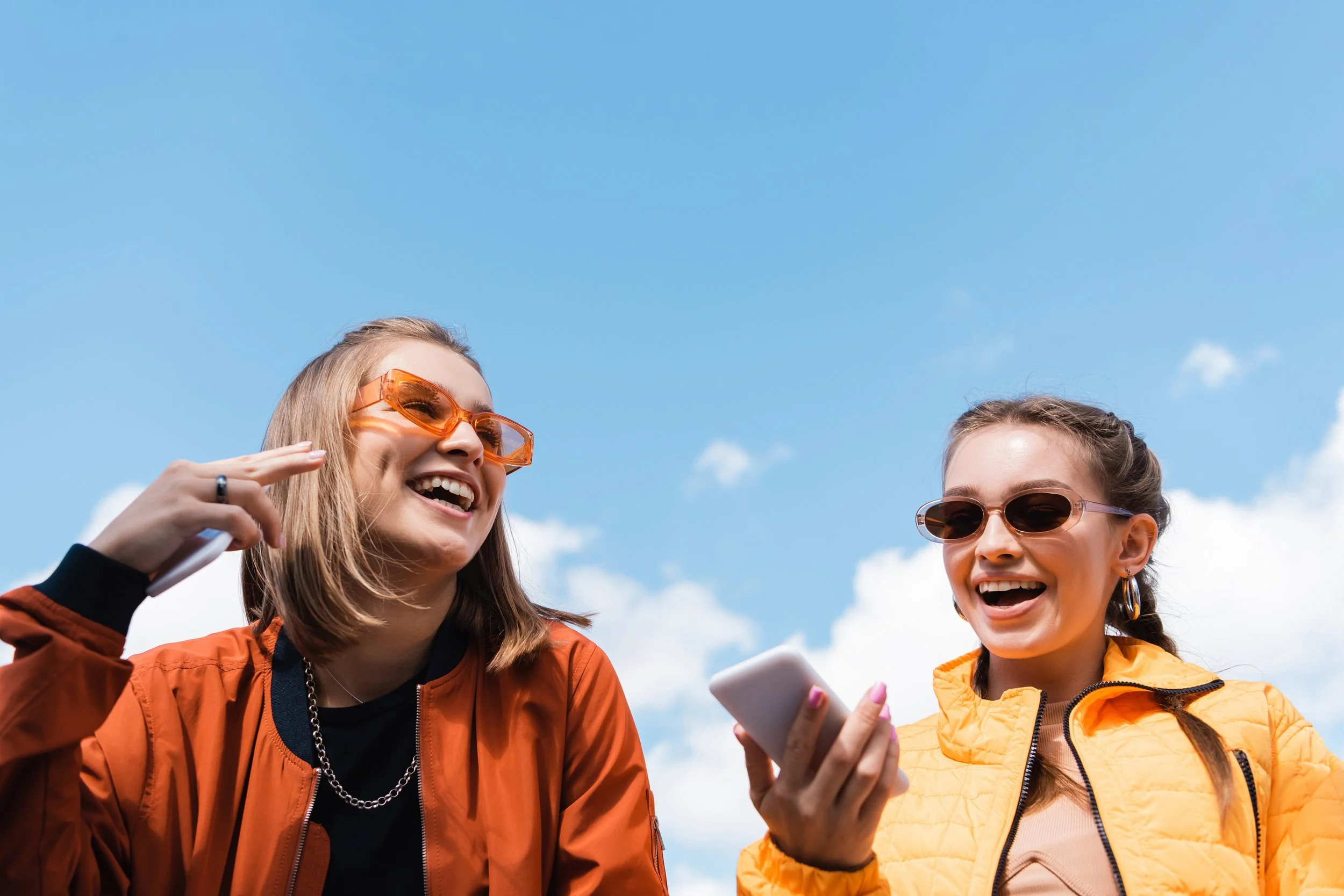 Two young women wearing sunglasses and orange jackets, laughing and talking outdoors under a blue sky.