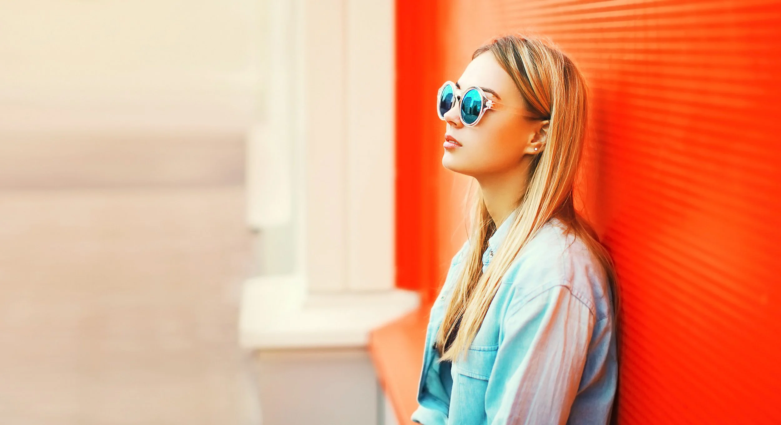 A young woman with long blonde hair wearing sunglasses with reflective blue lenses, dressed in a denim jacket, leaning against a bright orange wall, looking to the side.