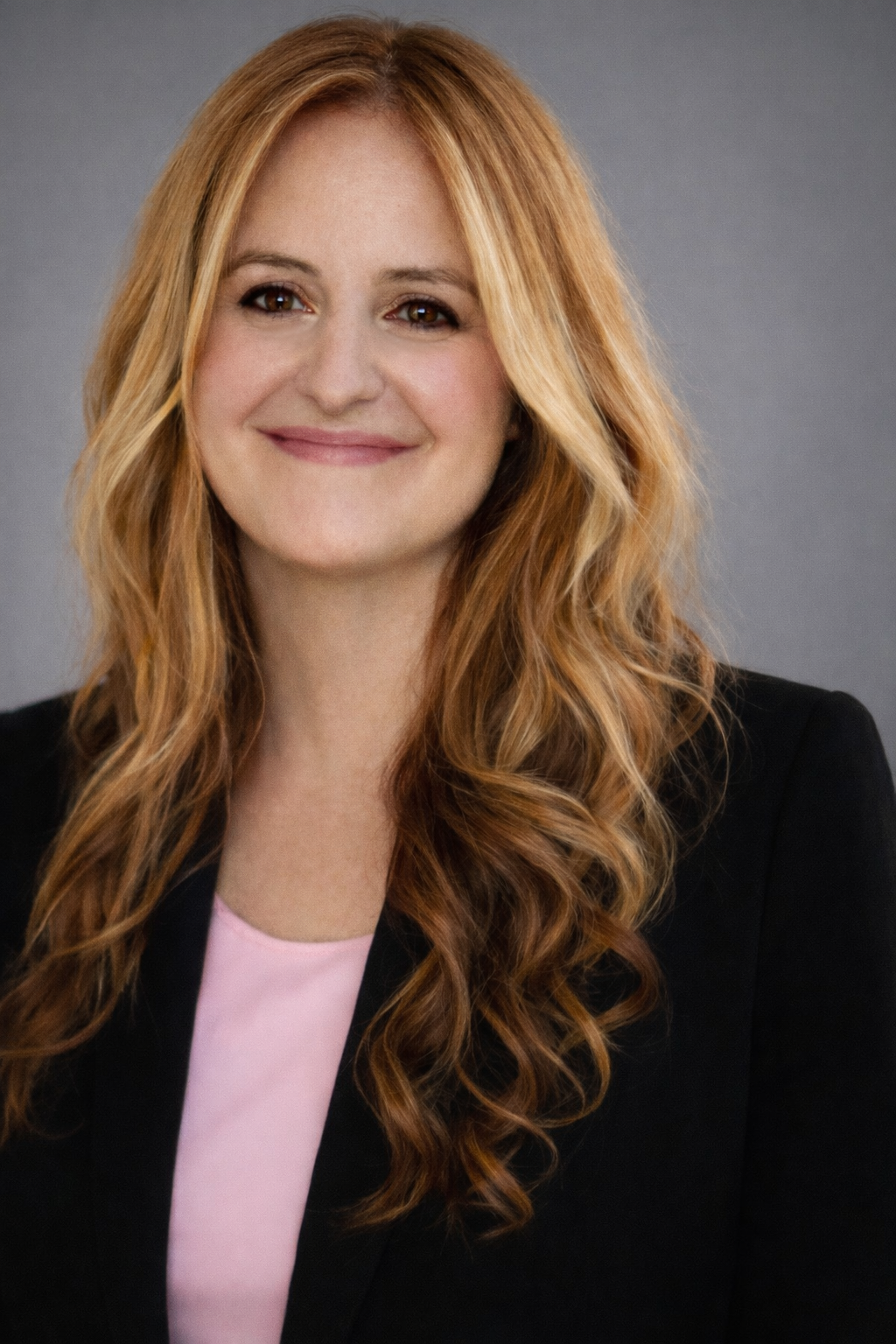 A woman with long, wavy red hair, wearing a pink top and black blazer, smiling against a gray background.
