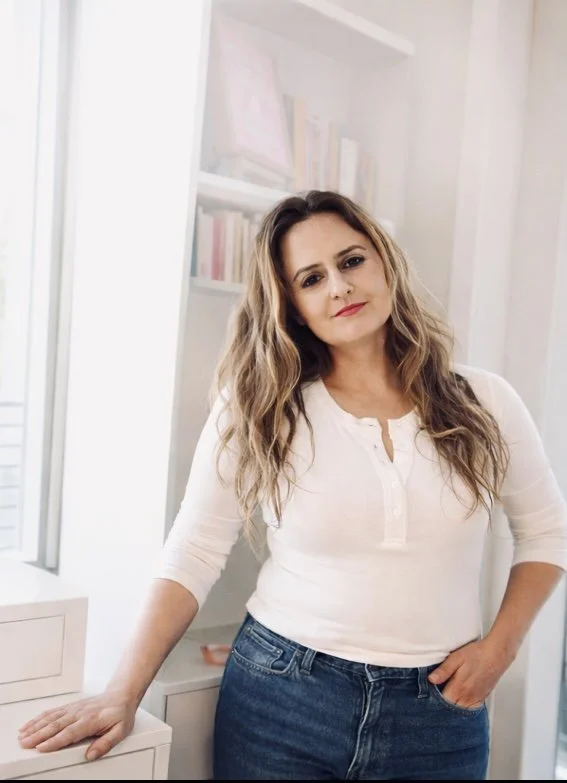 Woman with wavy, light brown hair wearing a white long-sleeve shirt and blue jeans, standing in a room with white shelves filled with books.