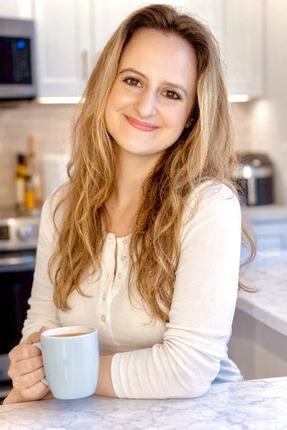 A woman with long, wavy hair is smiling while holding a blue mug, standing in a modern kitchen with white cabinets and appliances.