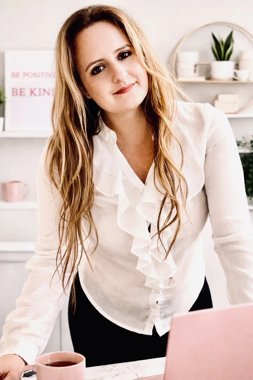 A woman with long, wavy, reddish-blonde hair and fair skin smiling at the camera, wearing a white blouse with ruffles, standing in a bright, modern kitchen with white shelves, potted plants, and a pink mug on the counter.