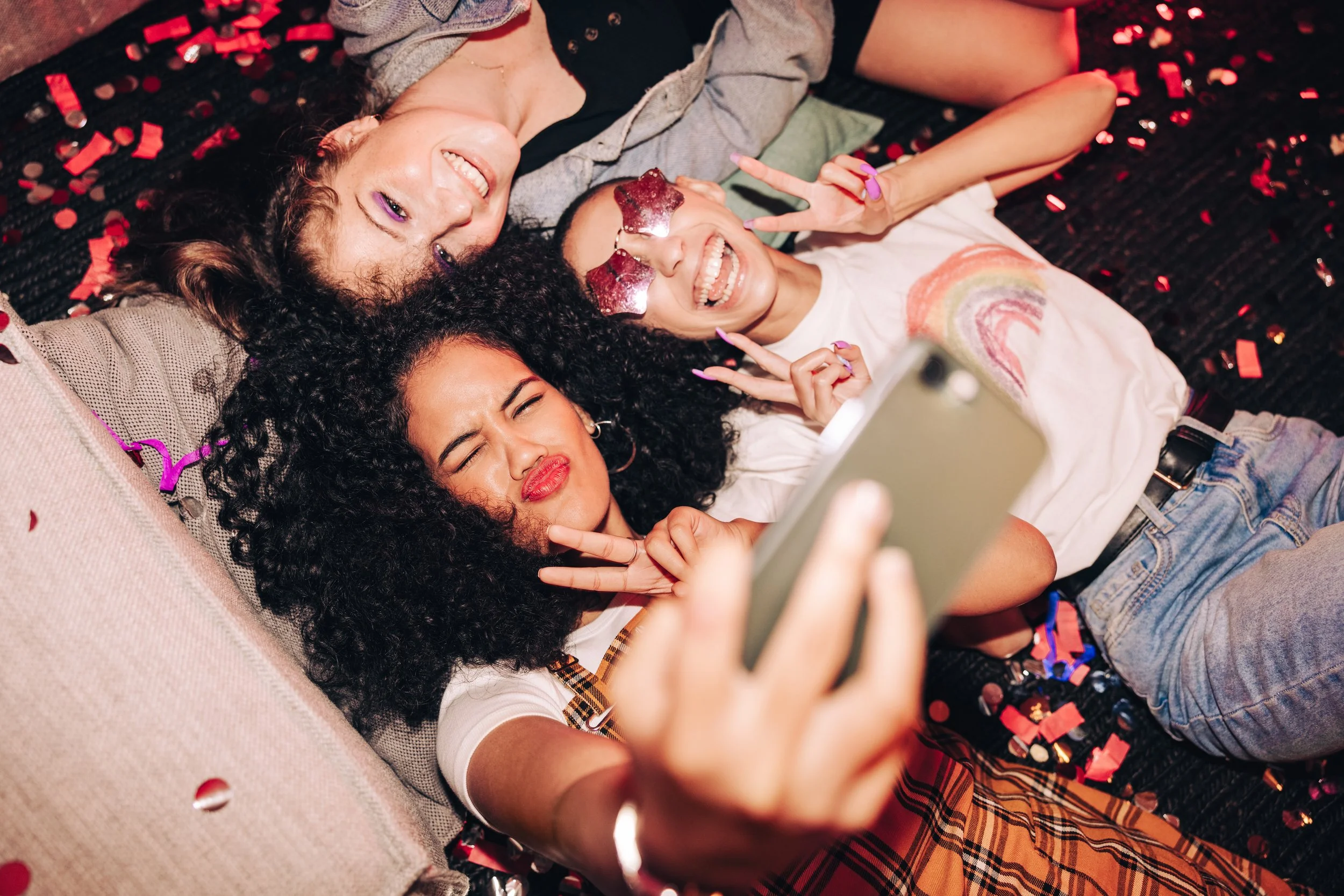 Three young women lying on a bed covered with colorful confetti, taking a selfie together. They are making playful facial expressions and peace signs, with happy and excited moods.