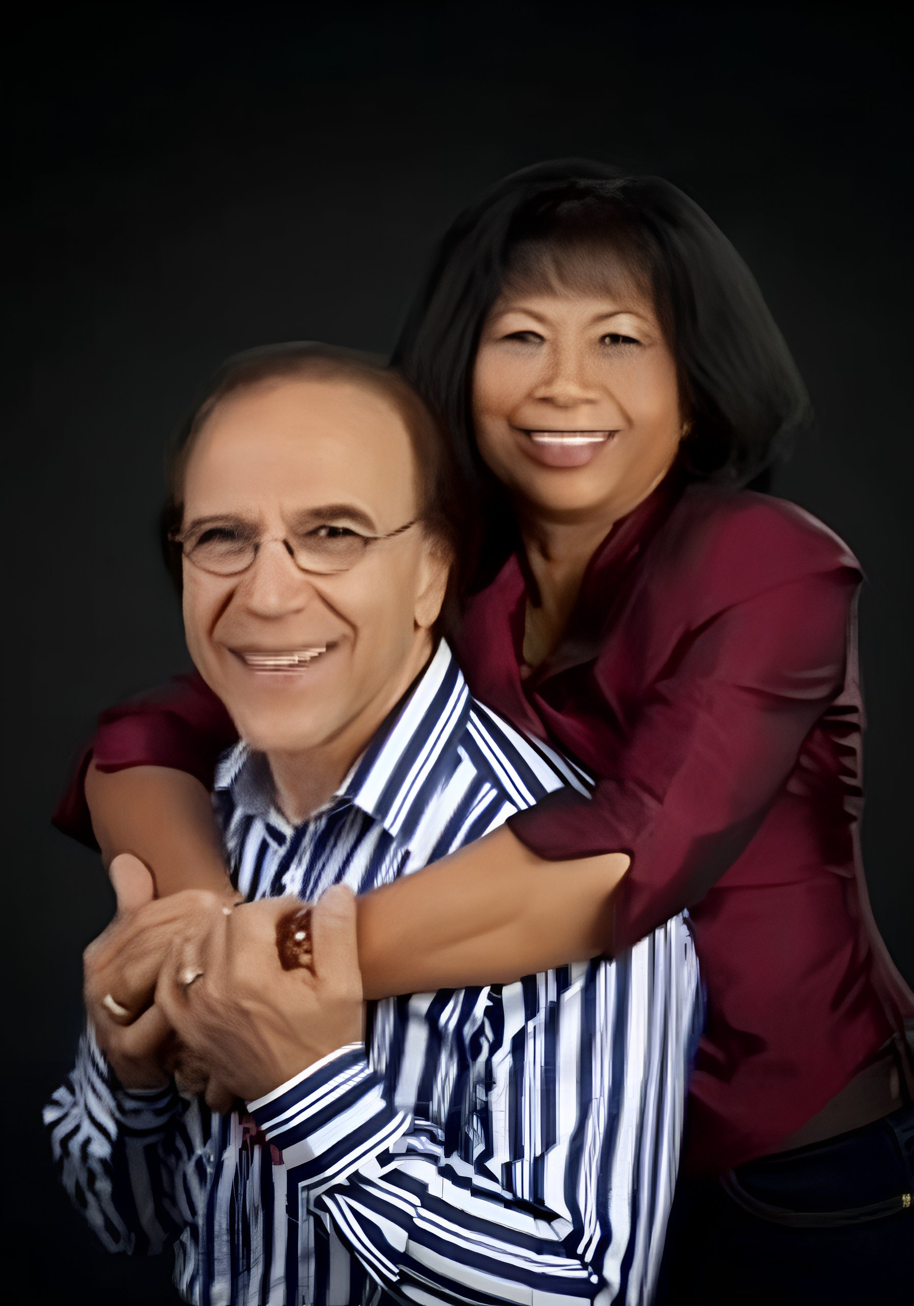 A smiling middle-aged man with glasses wearing a striped shirt, with a woman with black hair and a maroon blouse hugging him from behind against a black background.