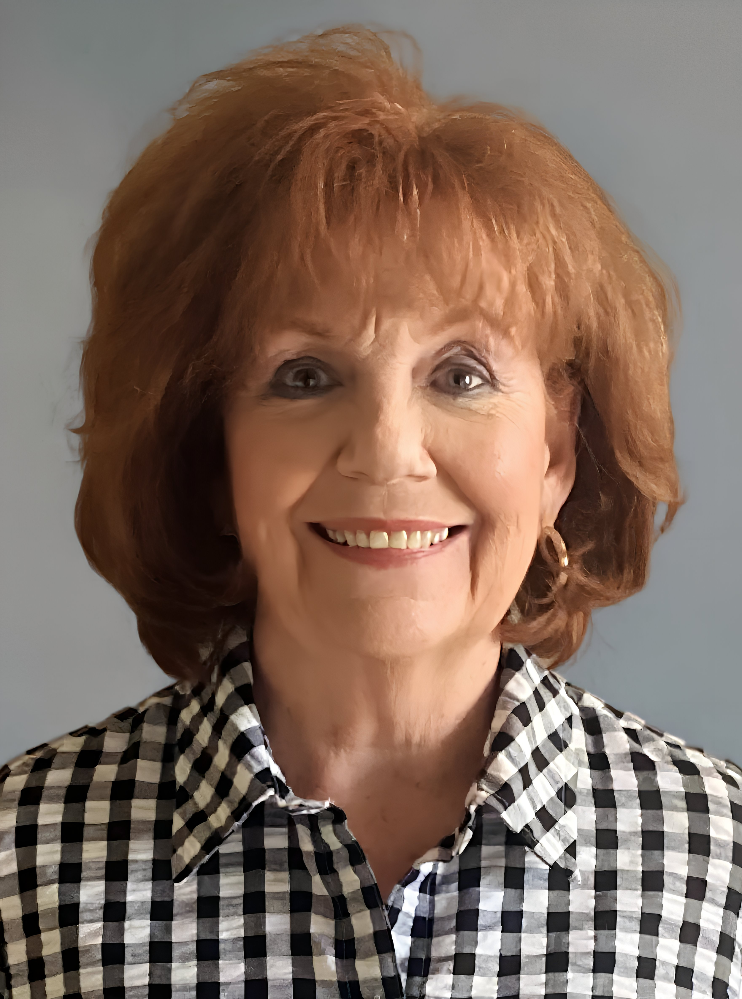 Portrait of a smiling middle-aged woman with curly red hair, wearing a black and white checkered shirt, against a plain background.