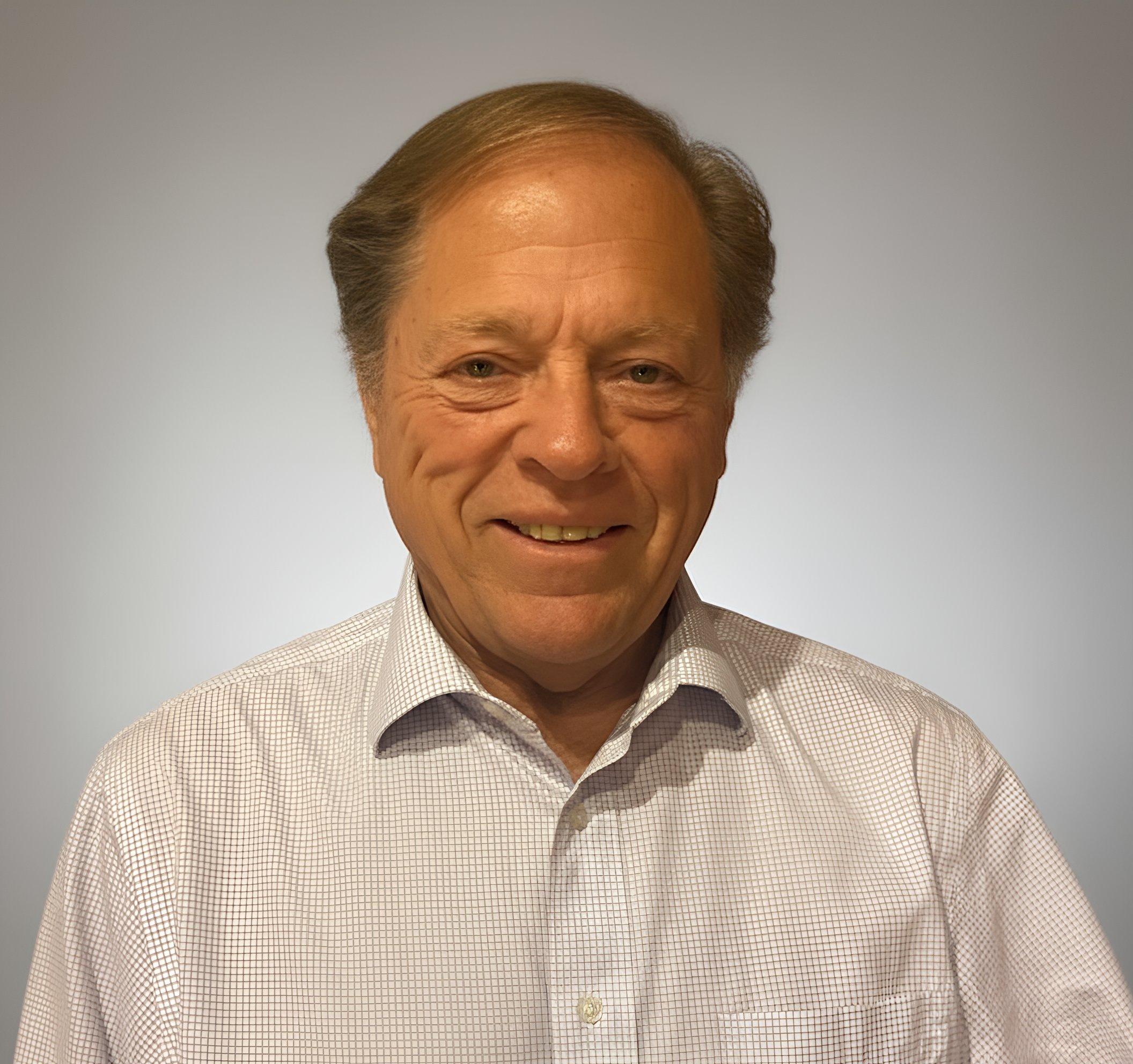 A middle-aged man with light skin, smiling, wearing a white checkered shirt, standing against a plain light gray background.
