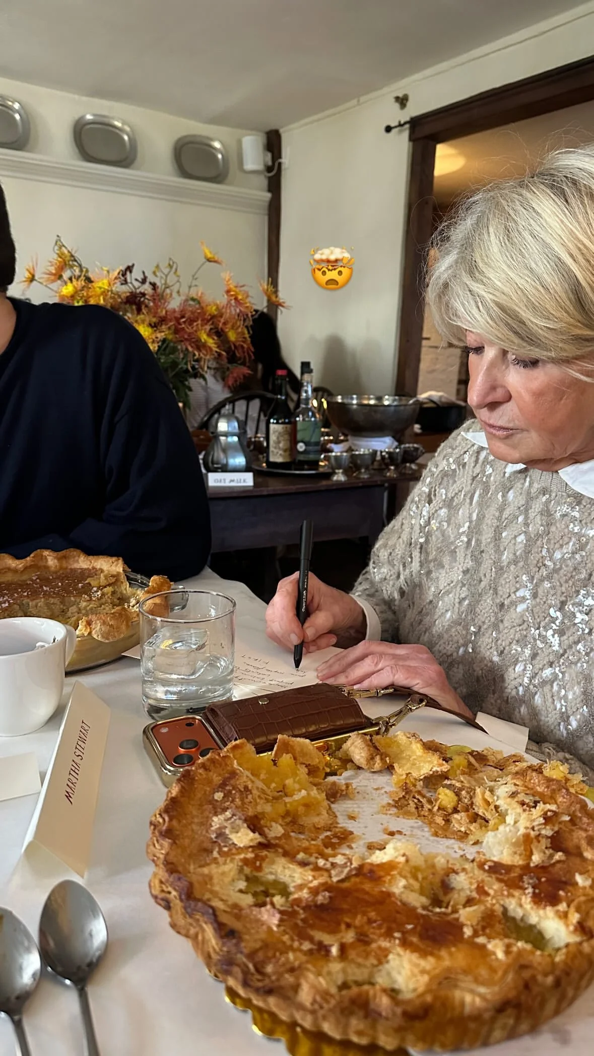 Martha Stewart, a woman with blonde hair in a shiny, patterned sweater, is writing notes at a table with a partially eaten pie, a cocktail, a white mug, and a brown wallet nearby. There are a floral arrangement and bottles in the background.