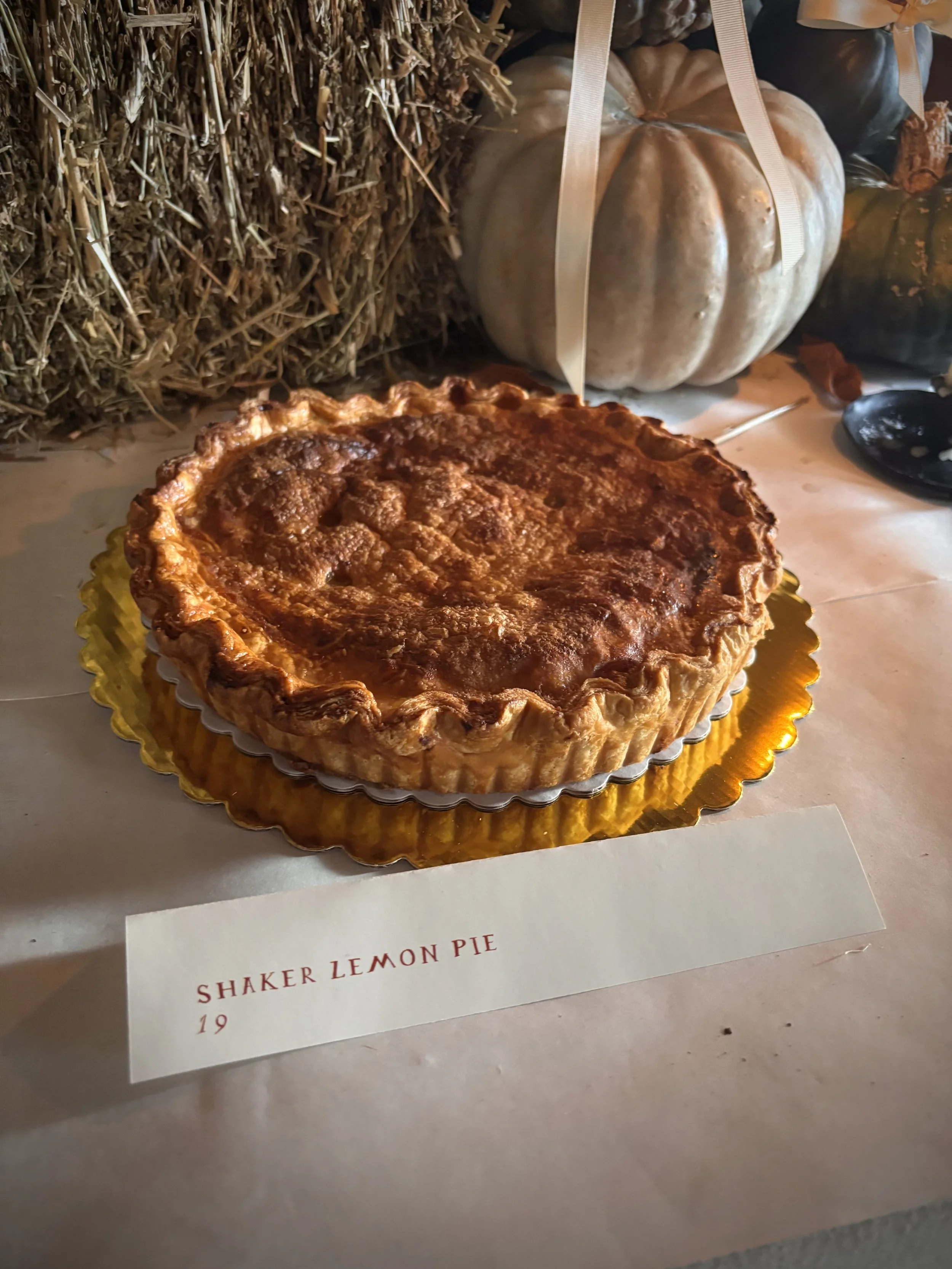 A pie with a golden crust labeled 'Shaker Lemon Pie' on a table with pumpkins and hay in the background.