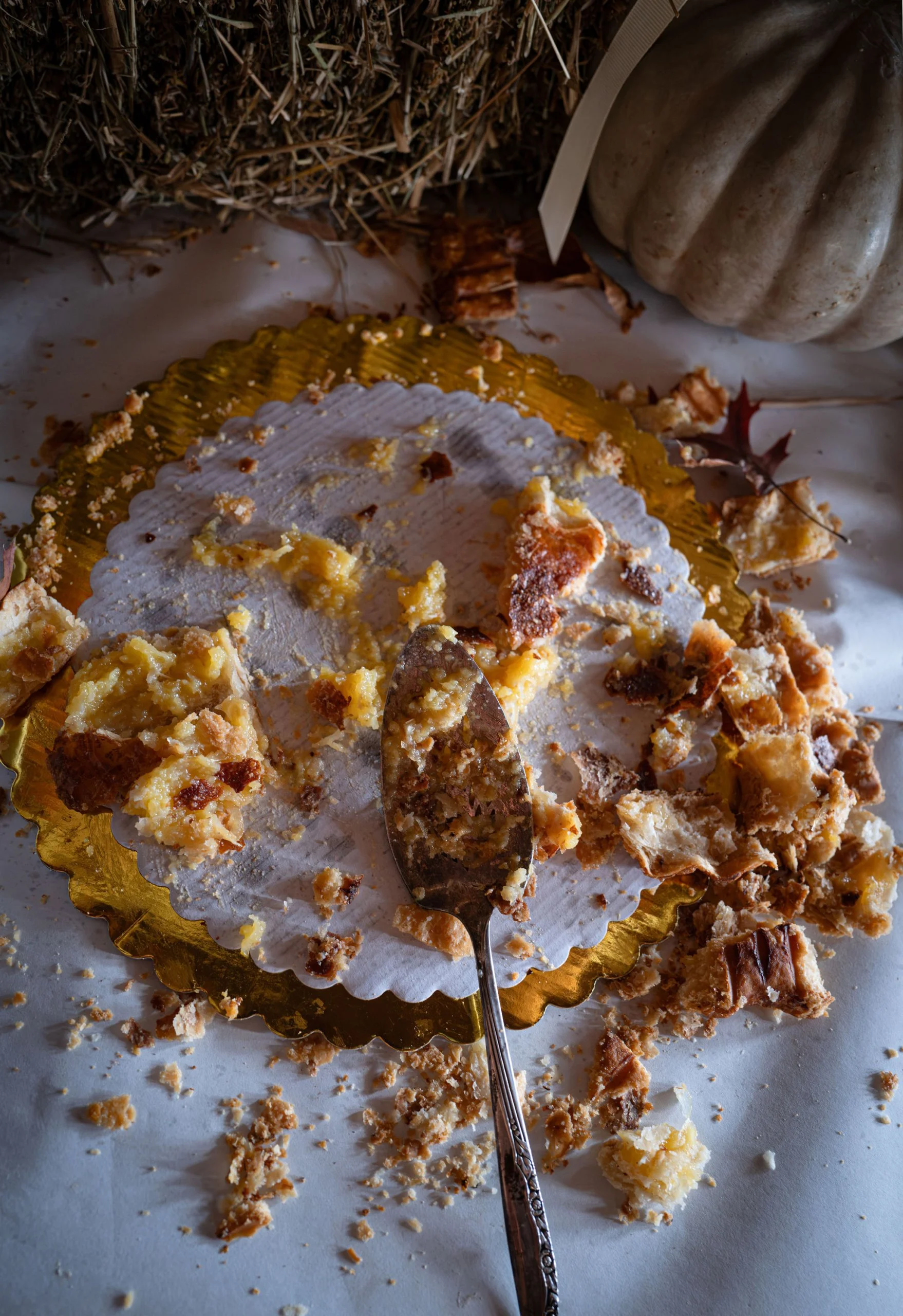 Close-up of a partially eaten shaker lemon pie on a cloth-covered table with crumbs scattered around, surrounded by autumn leaves and pumpkins.