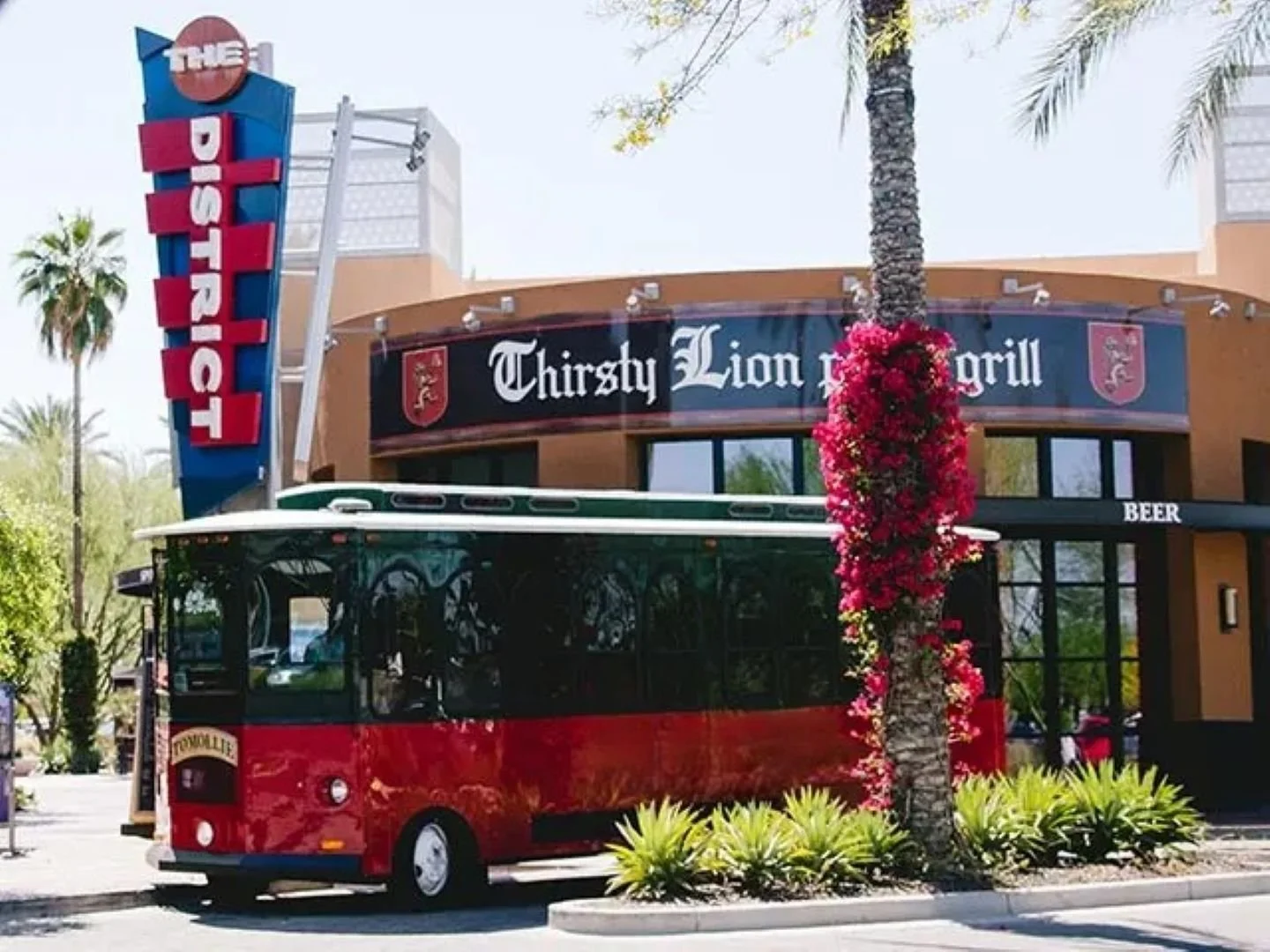 A red trolley parked in front of the Thirsty Lion Pub & Grill, with palm trees and flowering plants nearby.