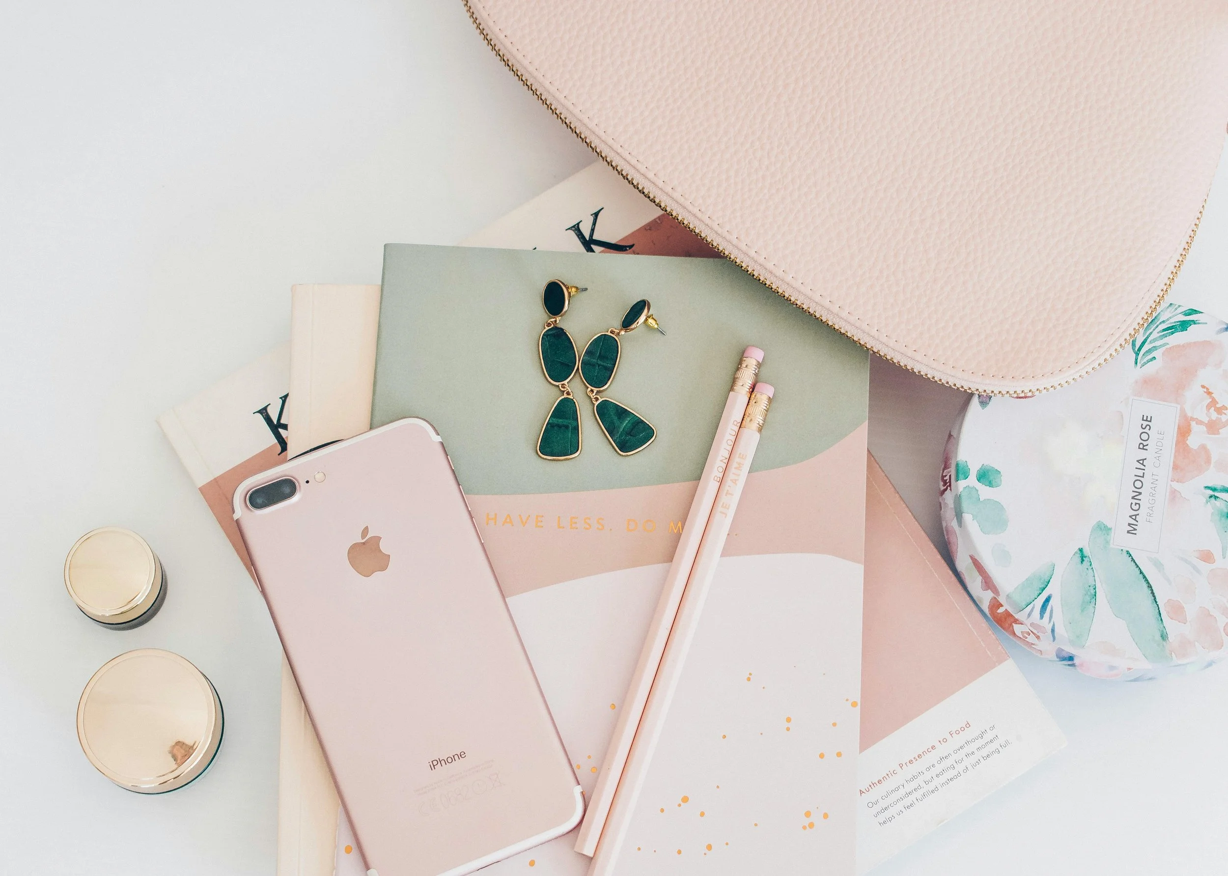 Flat lay of pink iPhone, gold and black earrings, pink pencils, a book, a large pink textured zippered pouch, a floral round container, and two small gold jars on a white surface.