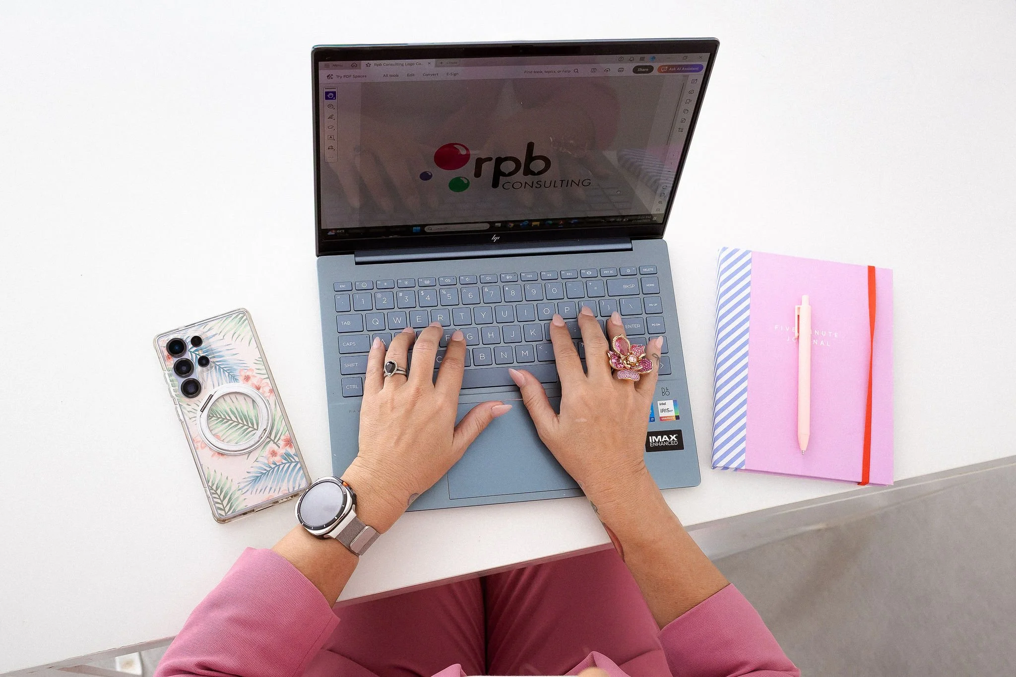 Top view of a person typing on a gray laptop with 'RPB Consulting' logo on the screen, placed on a white desk. The person wears a pink long-sleeve shirt, has various rings, a watch, and a bracelet. To the left, there is a phone with a tropical leaf case. To the right, a pink notebook with a pen on top.