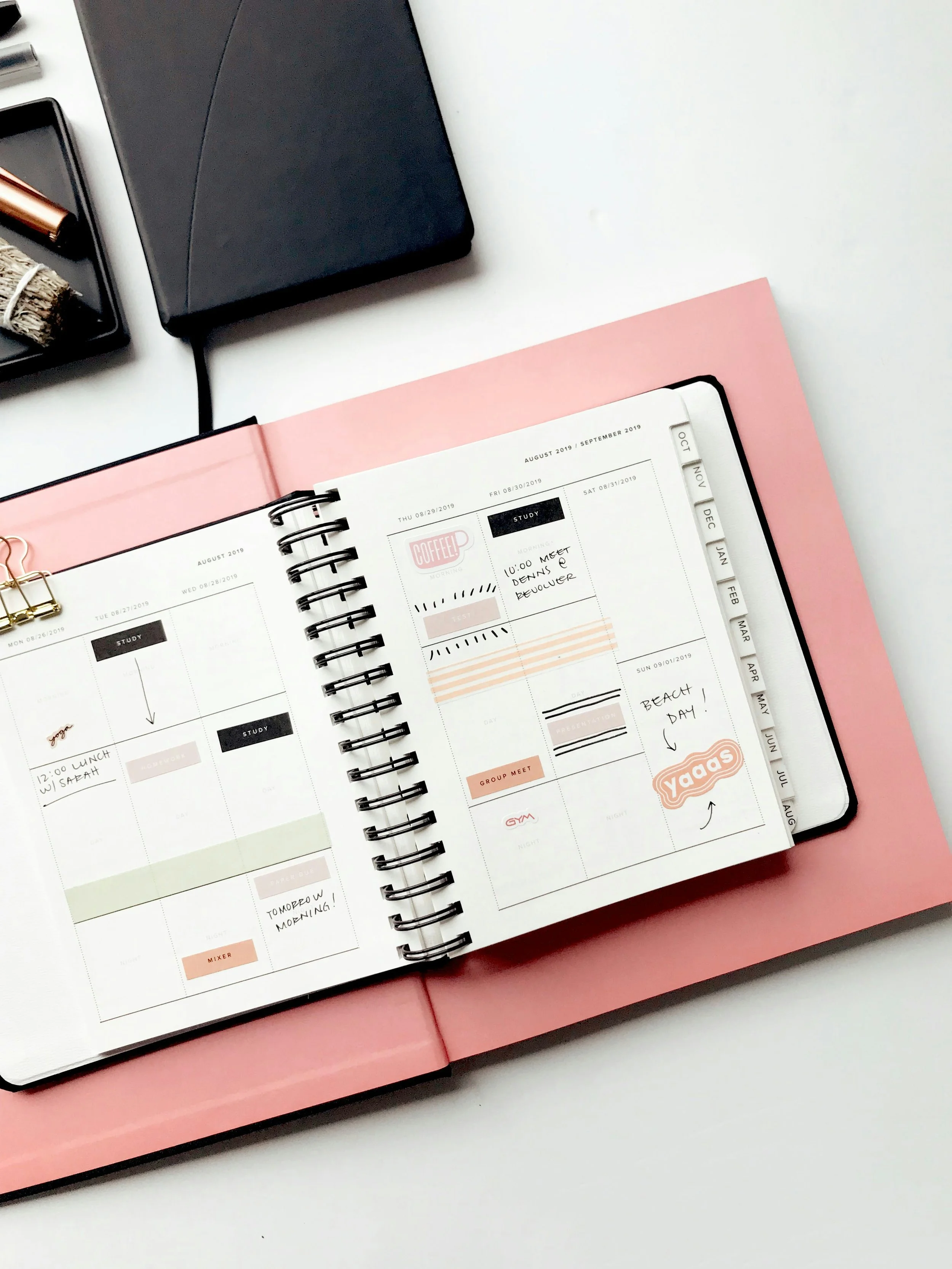 Open planner with handwritten notes and stickers, surrounded by a black notebook, a pink folder, and a small tray with miscellaneous items on a white surface.