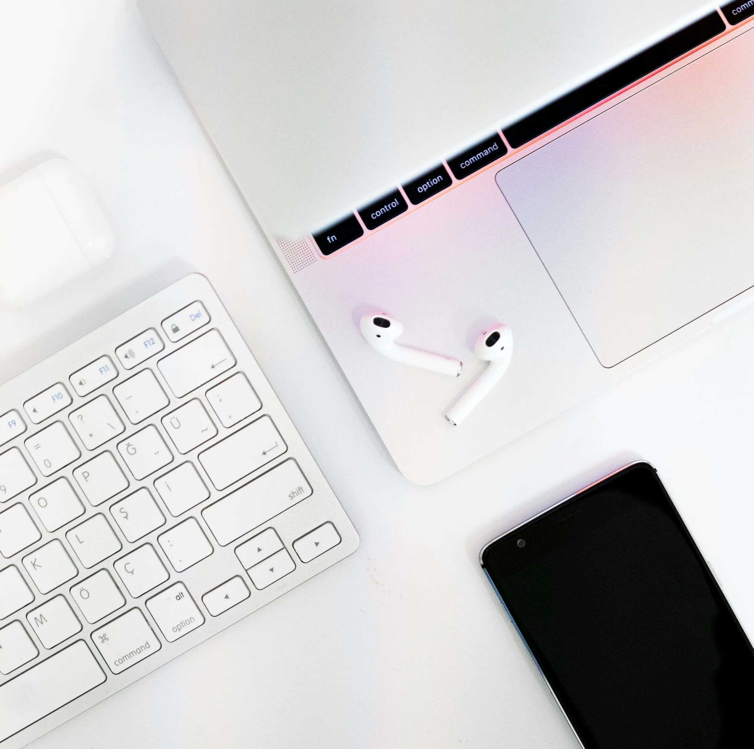 A flat lay of a white laptop, black and white wireless earbuds, a white keyboard, and a black smartphone on a white surface.