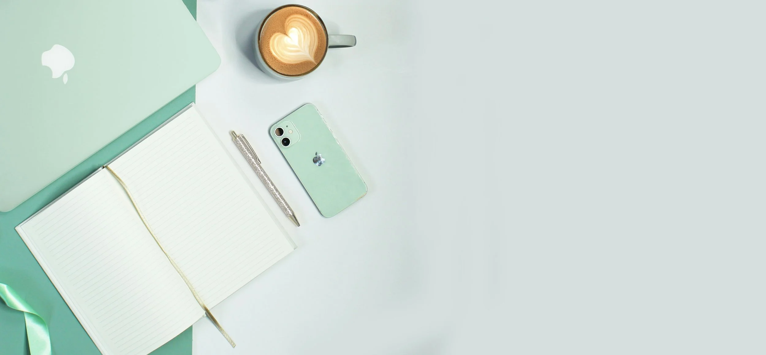 A flat lay of a workspace with a mint green laptop, a matching phone with a reflective apple logo, a silver glittery pen, an open lined notebook with a yellow ribbon bookmark, and a cup of coffee with heart-shaped latte art, all arranged on a white and mint green surface.