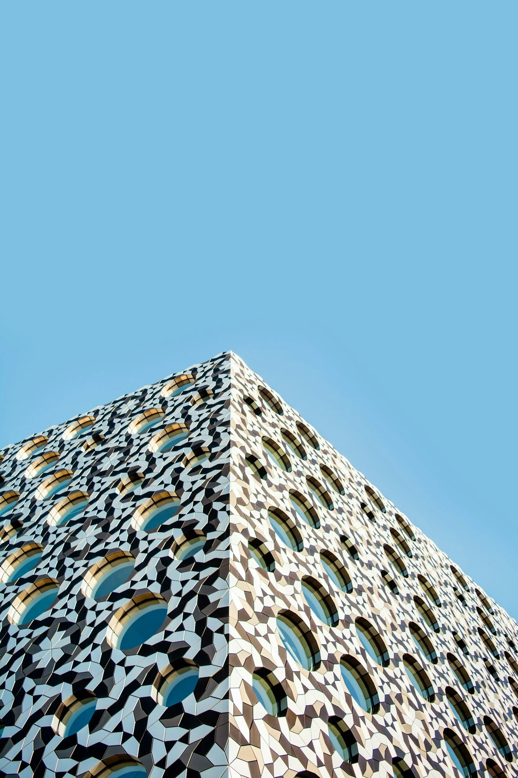 A modern building with a geometric patterned facade and numerous circular windows, set against a clear blue sky.
