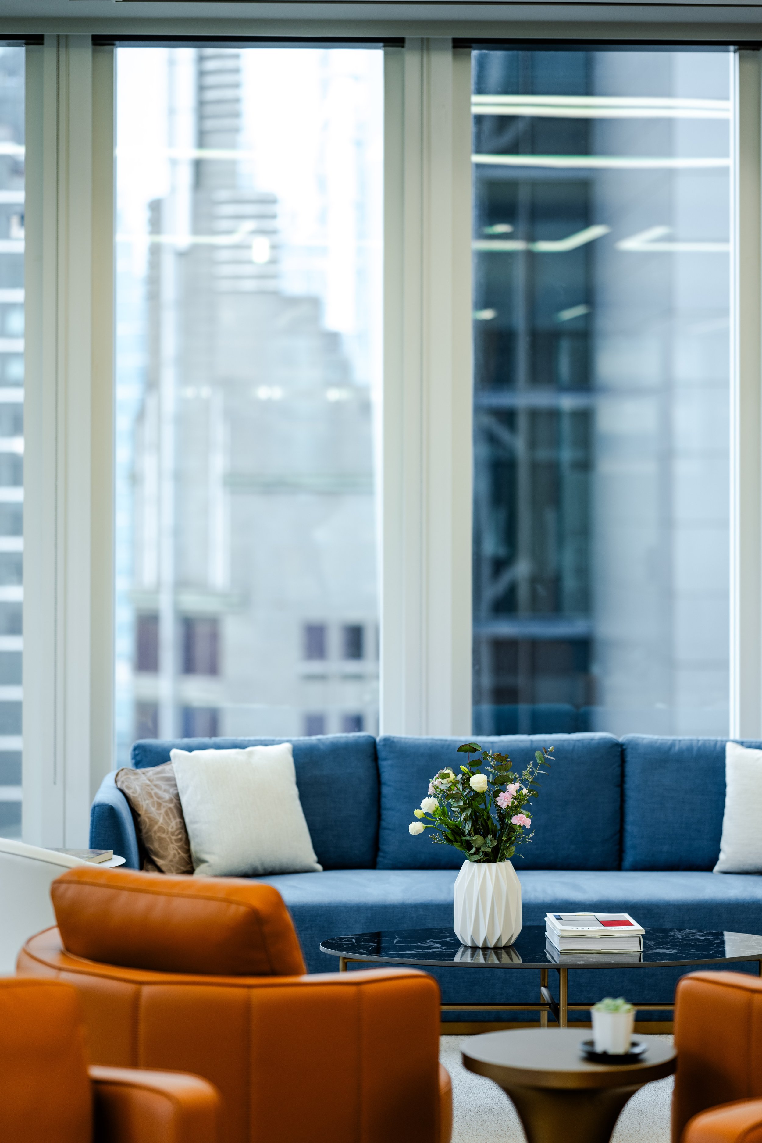 Interior of a modern living room with a large window, a blue sofa with throw pillows, a black coffee table with a white vase and flowers, and orange chairs.
