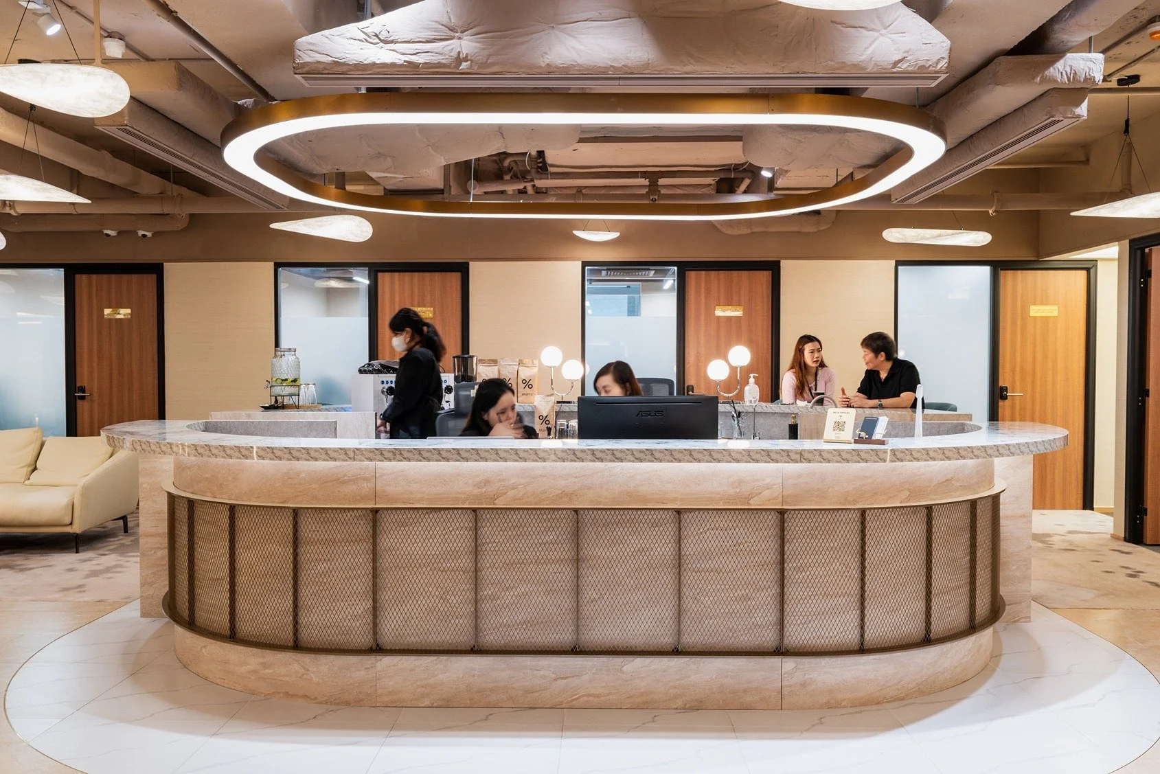Modern hotel check-in counter with three staff members attending to guests, surrounded by seating area and doors in the background.