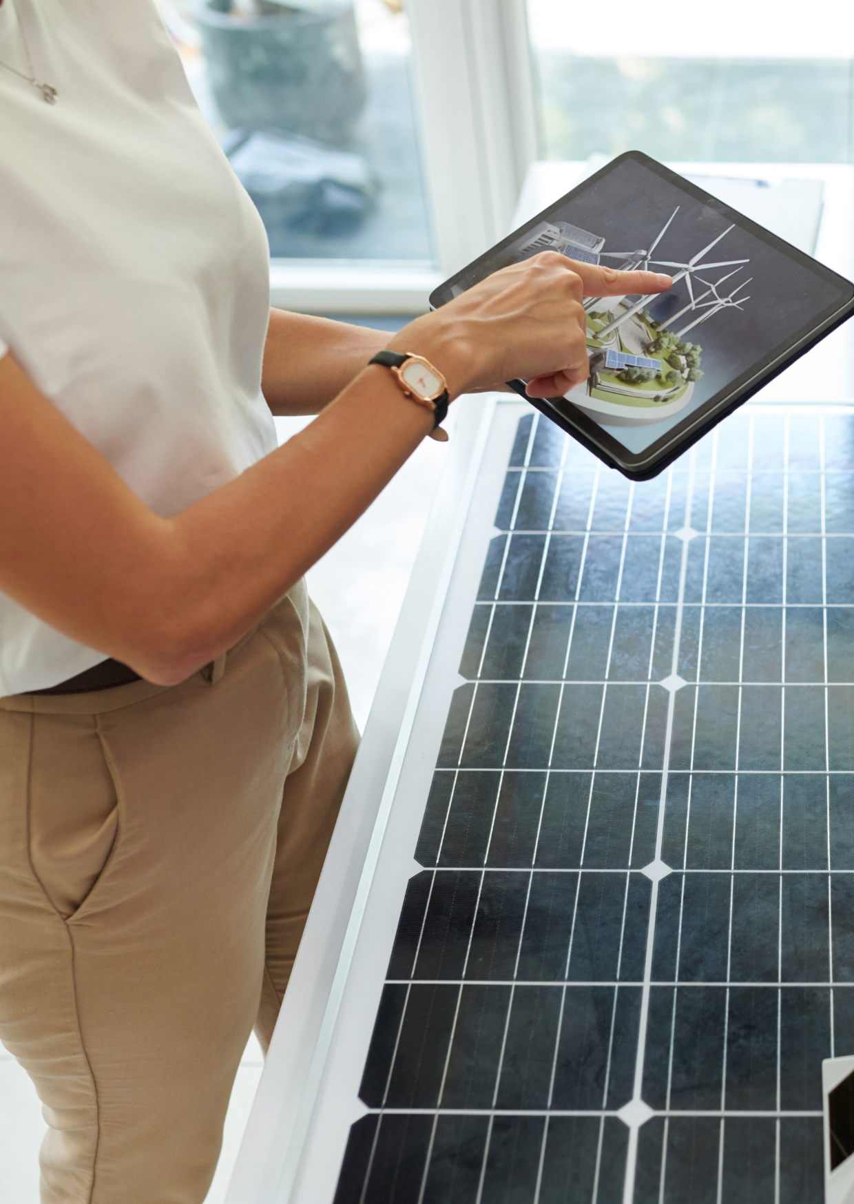 Person using a digital tablet with a 3D rendering of wind turbines, solar panels, and a power station, standing next to a solar panel on a table near a window.