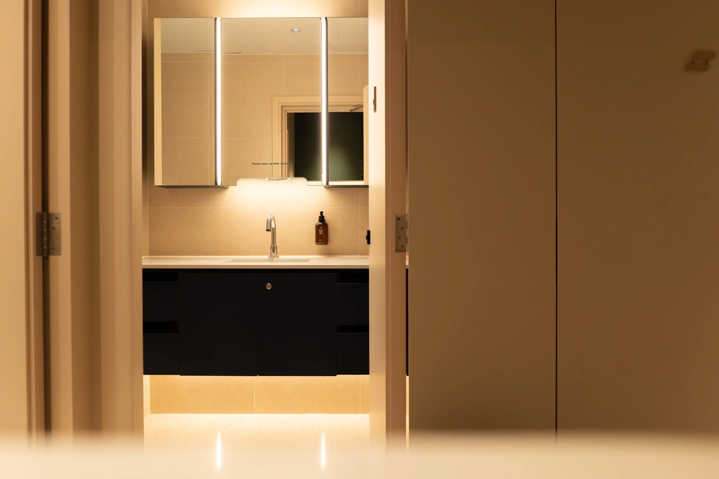 A modern bathroom seen through a partially open door. The bathroom features a black vanity with a sink, a sleek faucet, a brown soap dispenser, and a wall-mounted mirror with built-in lighting. The wall is tiled in light neutral tones, and the room has contemporary, minimalist decor.
