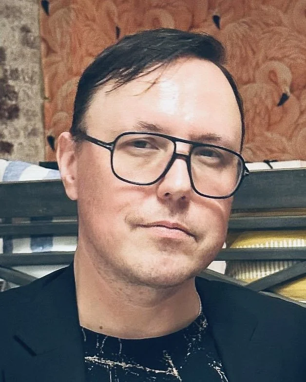 A man with short dark hair and glasses, wearing a black blazer over a black shirt with white paint splatter design, in an indoor setting with a patterned wallpaper background and shelves with plates.
