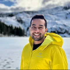 A smiling man in a yellow jacket standing in a snowy landscape with mountains in the background.