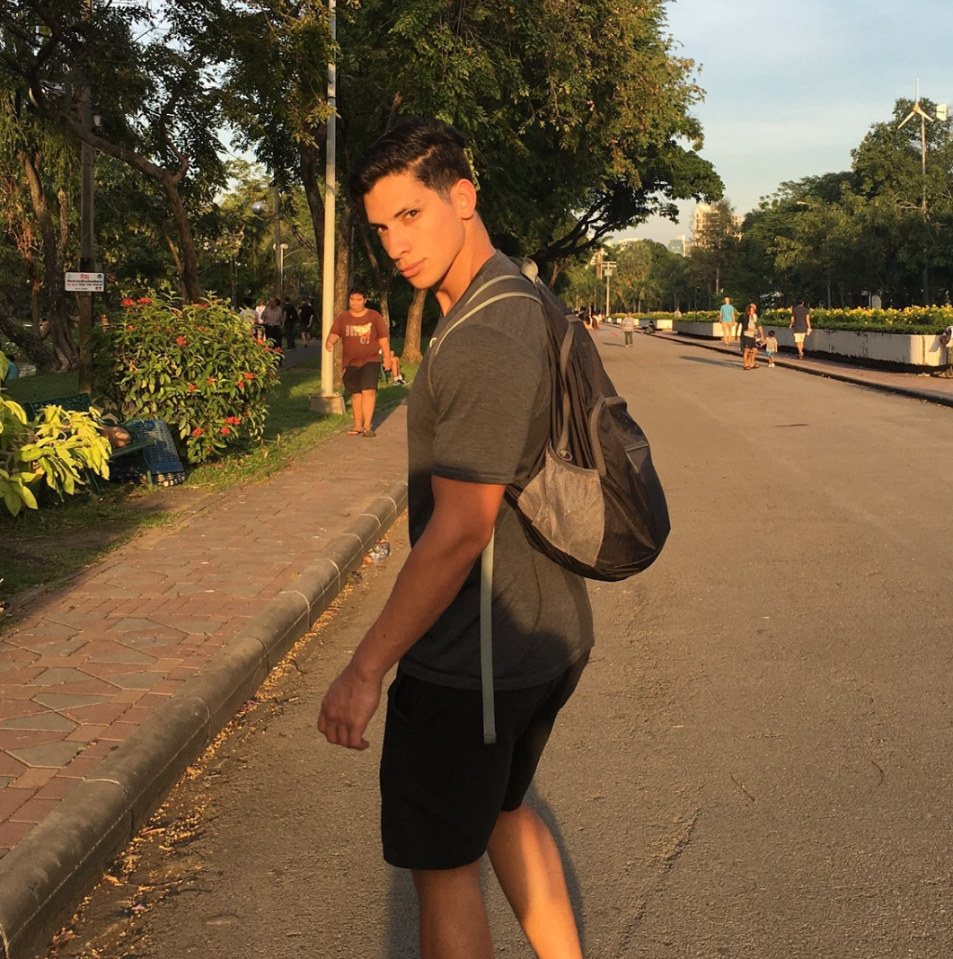 Young man with dark hair, wearing a gray t-shirt and black shorts, stands on a city sidewalk with a backpack, in a park-like area with trees and other pedestrians during sunset.
