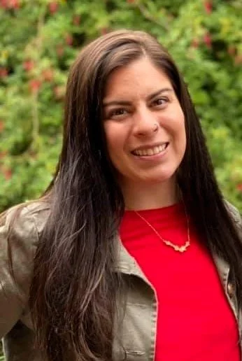 A smiling woman with shoulder-length black hair wearing a blue top, standing outdoors with green plants in the background.