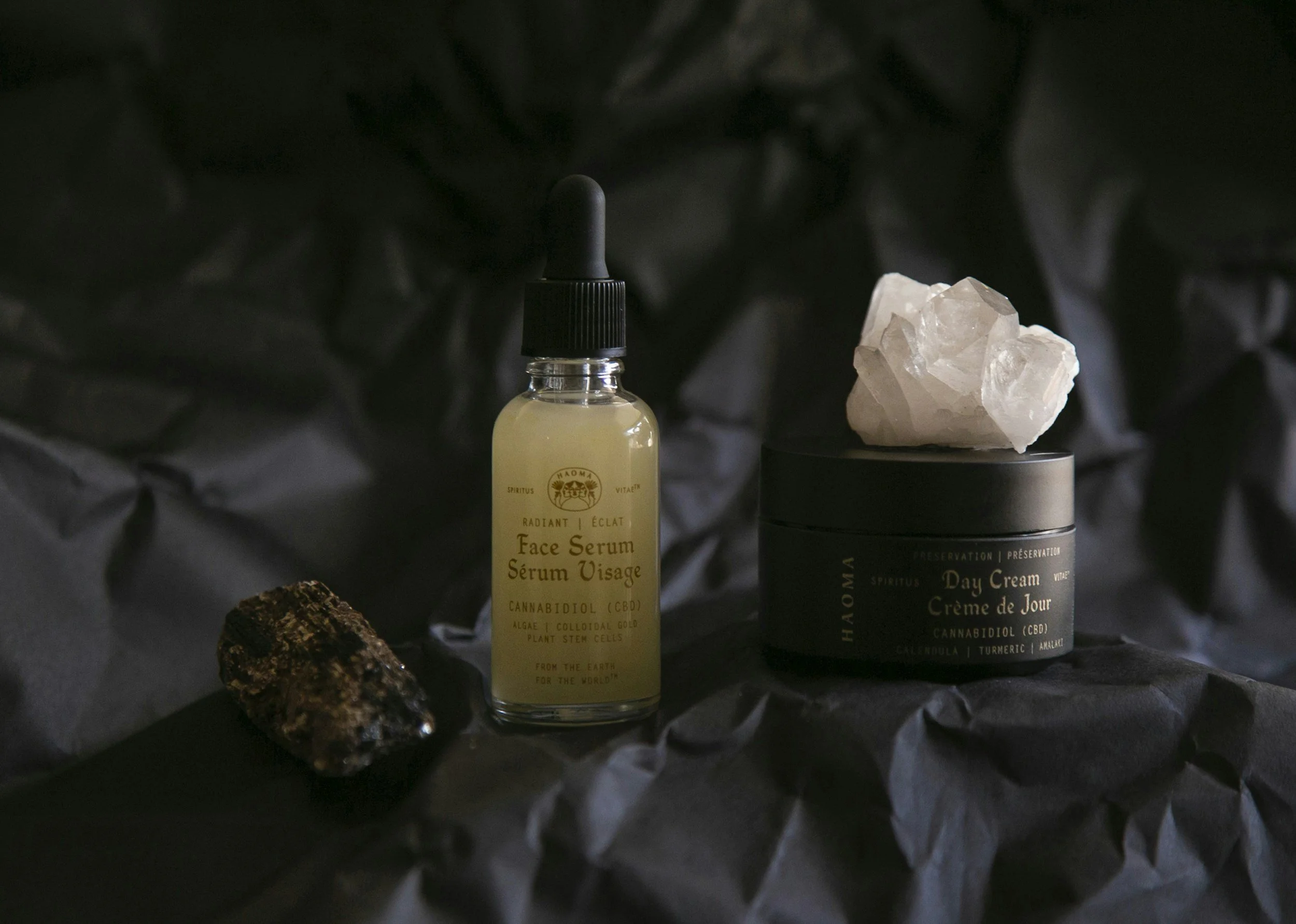 A small bottle of face serum, a container of day cream with a white mineral on top, a rock, all placed on crumpled black paper.